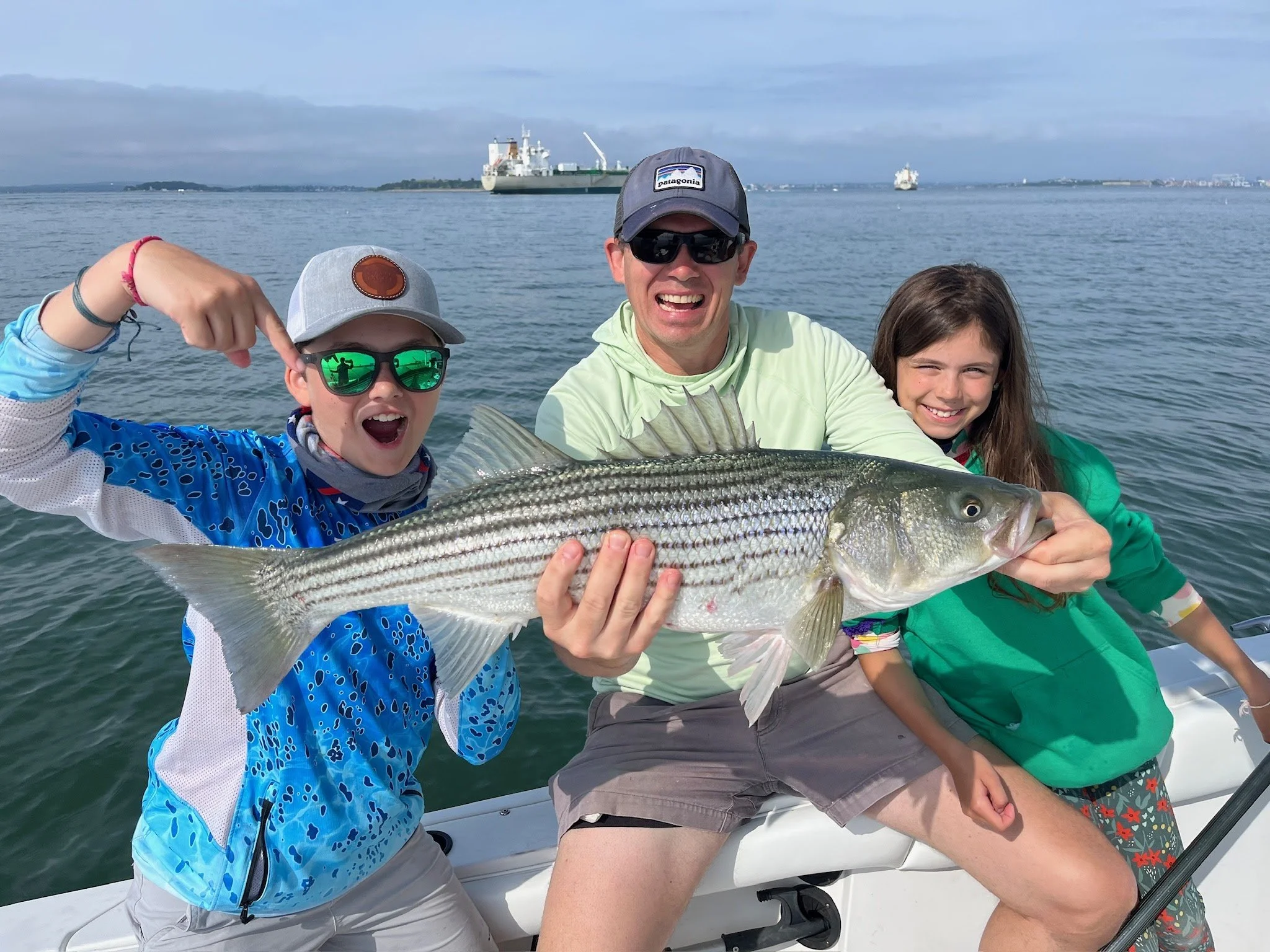 Rob Wittmann and his children on a boat holding a large striped bass they caught while fishing in the water. They are smiling and enjoying a day on the water with ships in the background.