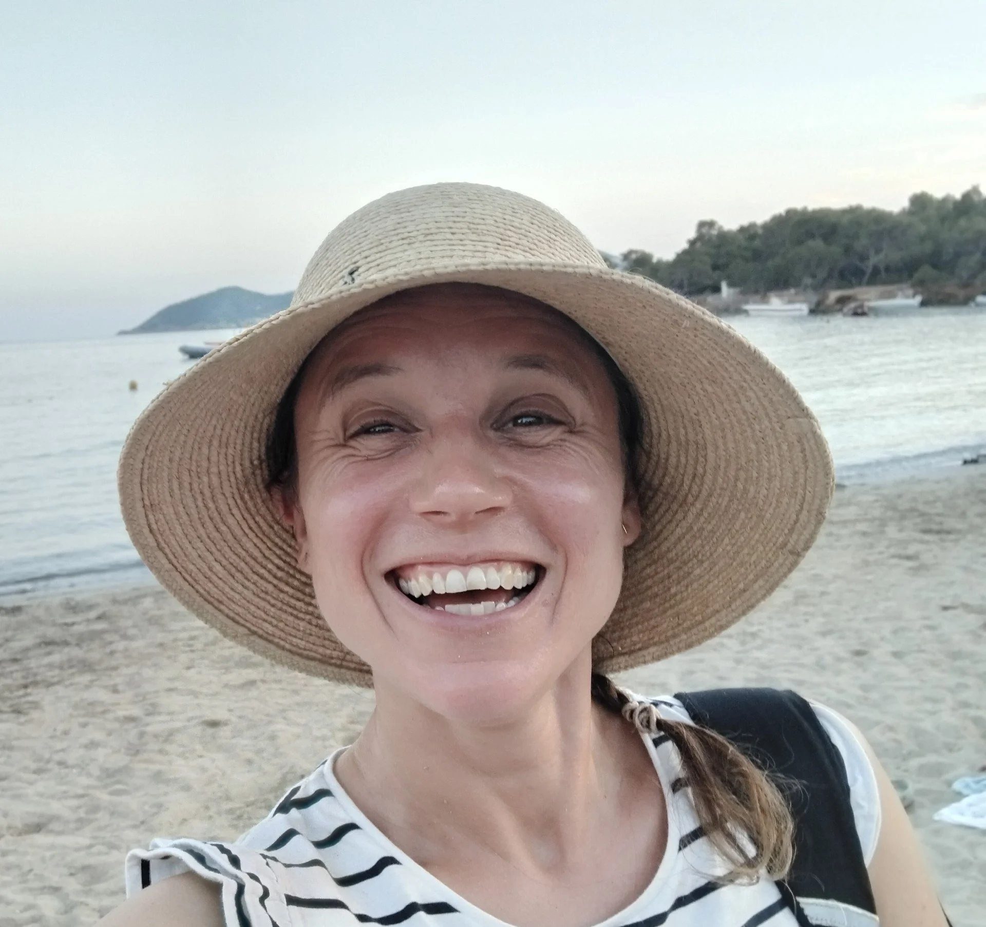 Woman in a striped shirt and large sun hat smiling on the beach with water, boats, and trees in the background.