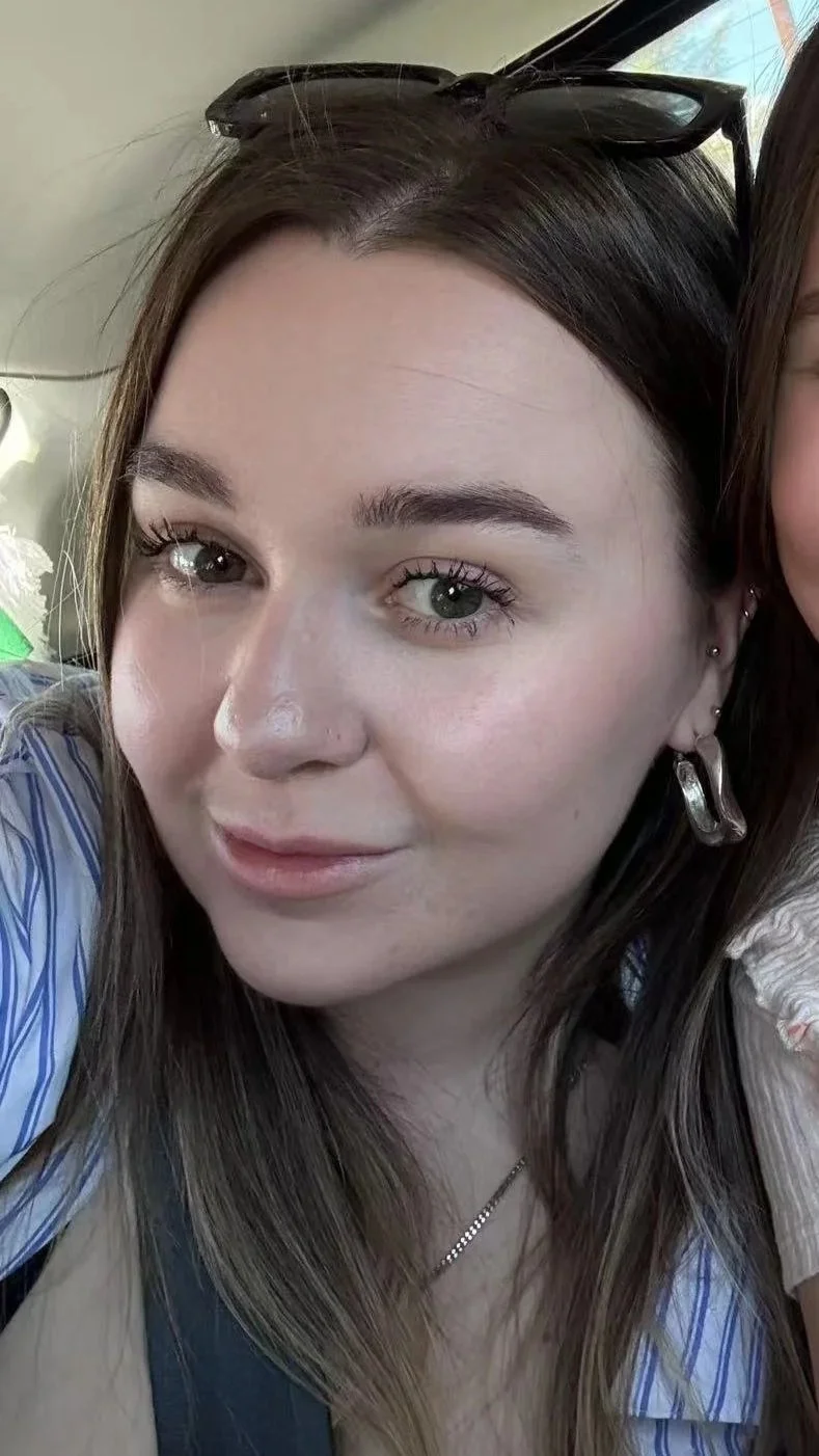 A young woman with long brown hair, blue eyes, and light skin, wearing sunglasses on her head, earrings, and a striped shirt. She is smiling softly and looking at the camera.