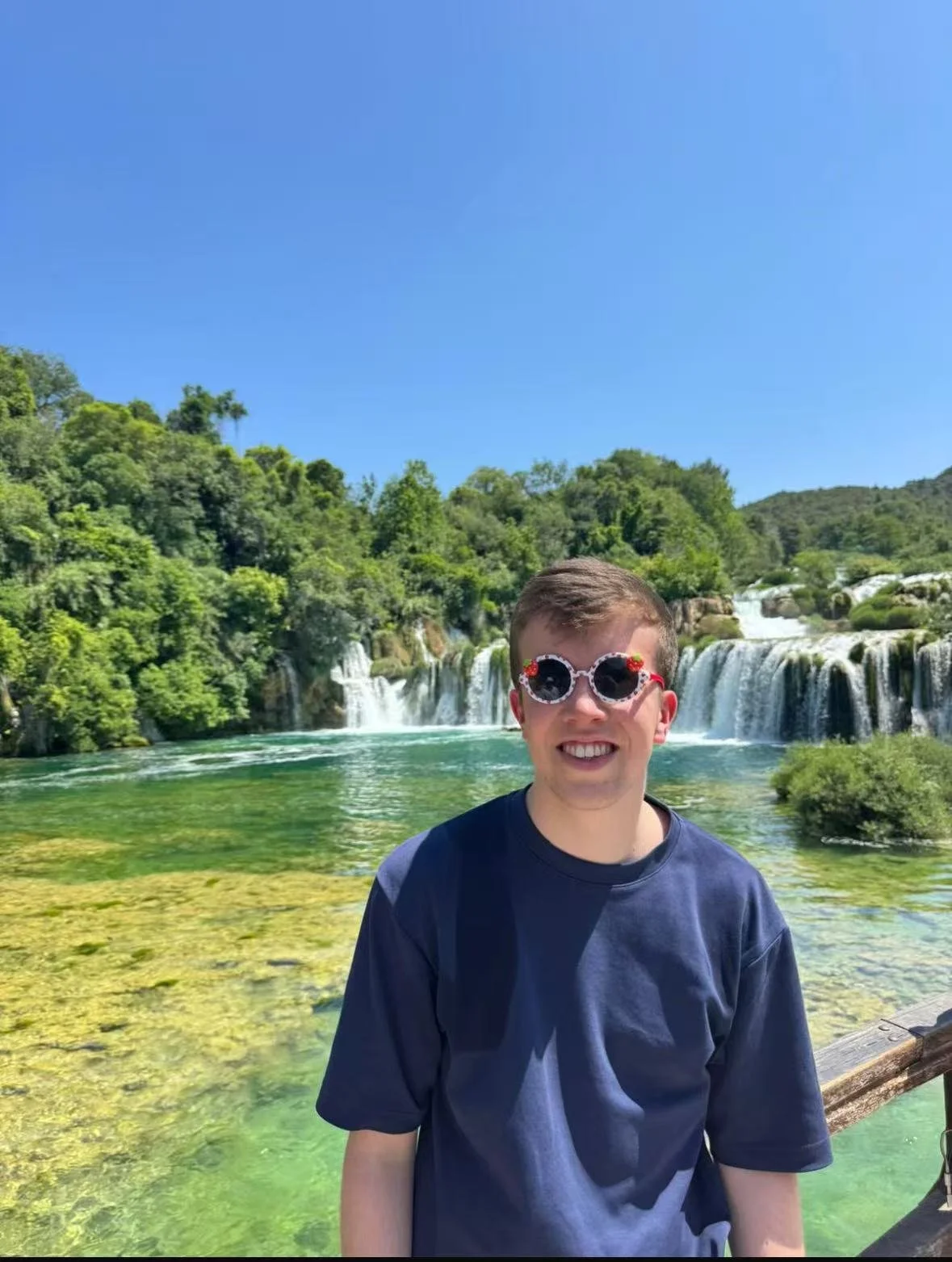 A young man with sunglasses and a navy shirt standing in front of a waterfall and lush green trees under a clear blue sky.