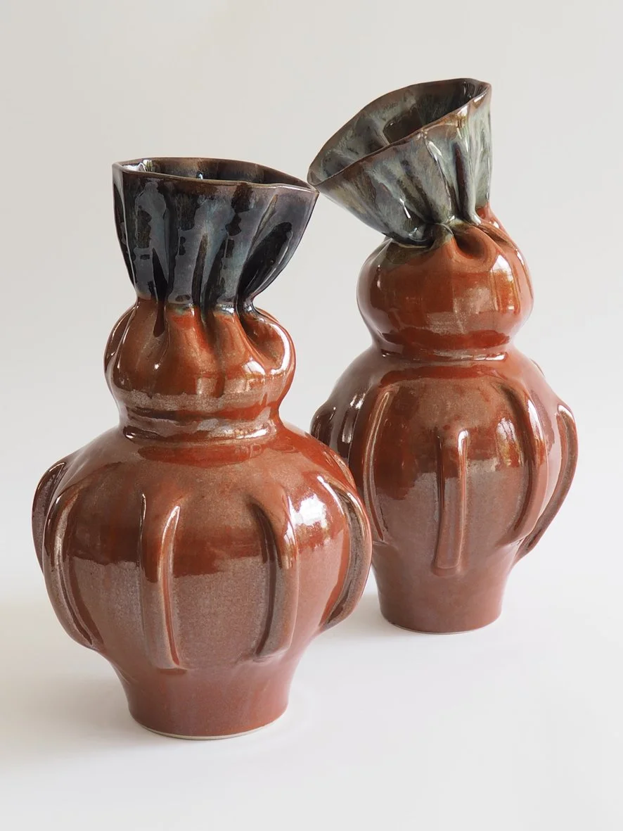 Two ceramic vases with rounded bodies and flared tops, glazed in reddish-brown with dark accents, positioned on a white surface against a plain background.