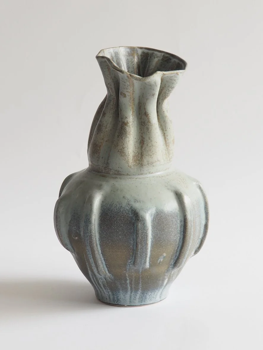 A ceramic vase with a unique, irregular shape and a textured, glazed surface in shades of white, gray, and brown, set against a plain background.