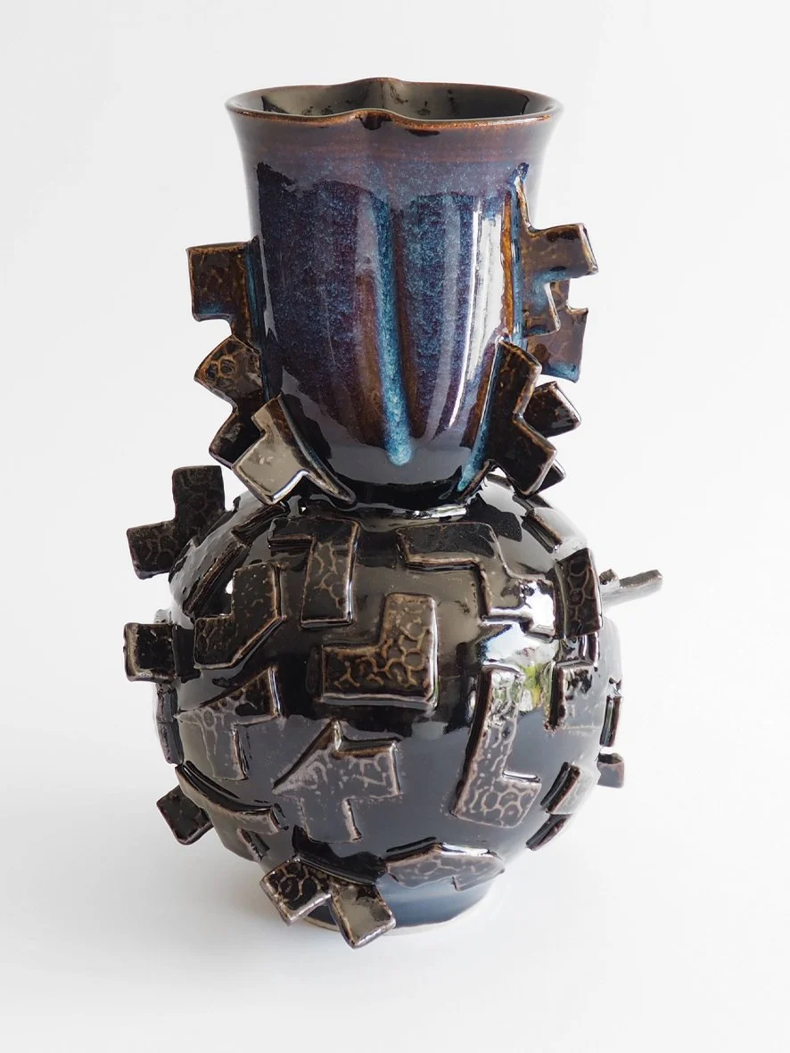 A decorative ceramic vase with a glossy dark glaze, featuring geometric protrusions around the body and a flared opening at the top, set against a plain white background.