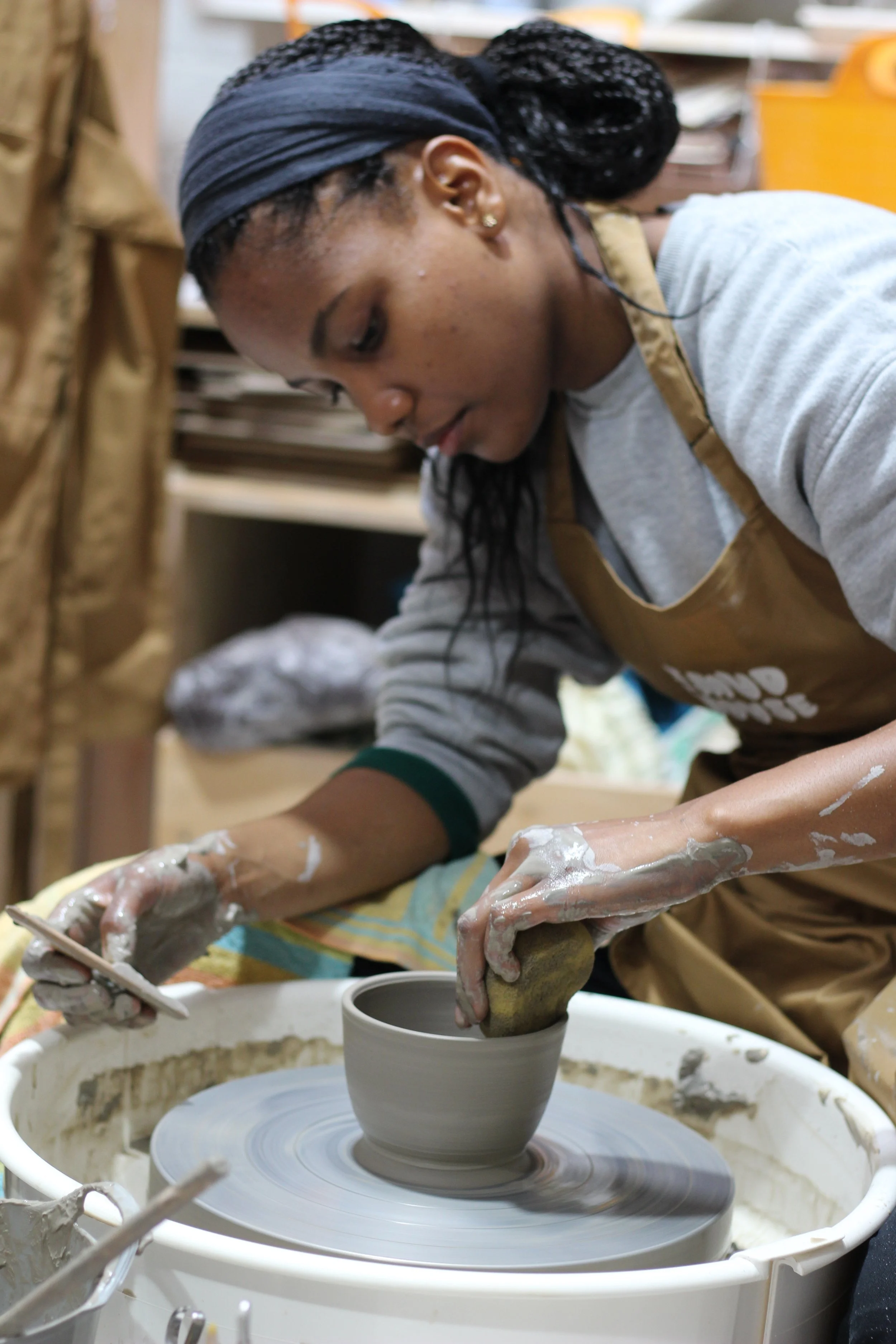 A young woman using a pottery wheel to shape a clay bowl in a pottery studio, with her hands covered in clay.