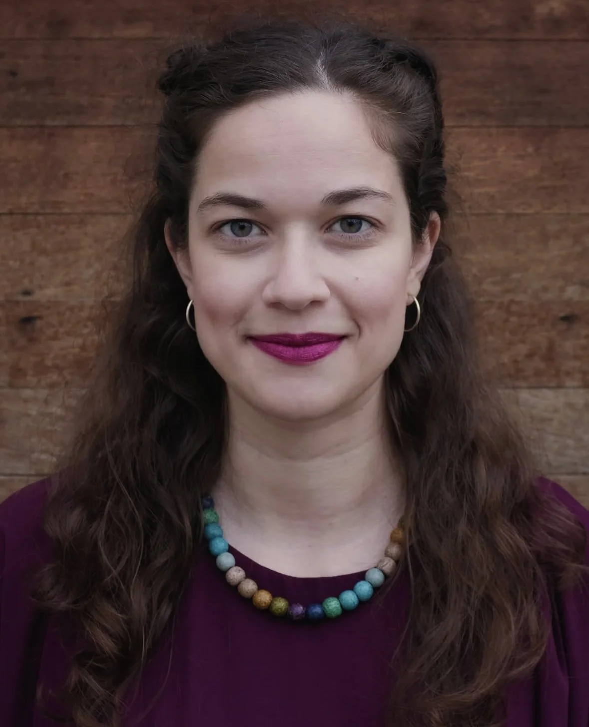 A woman with long, wavy brown hair, blue eyes, and pink lipstick, wearing a purple top, a multicolored beaded necklace, gold hoop earrings, and standing in front of a wooden background.
