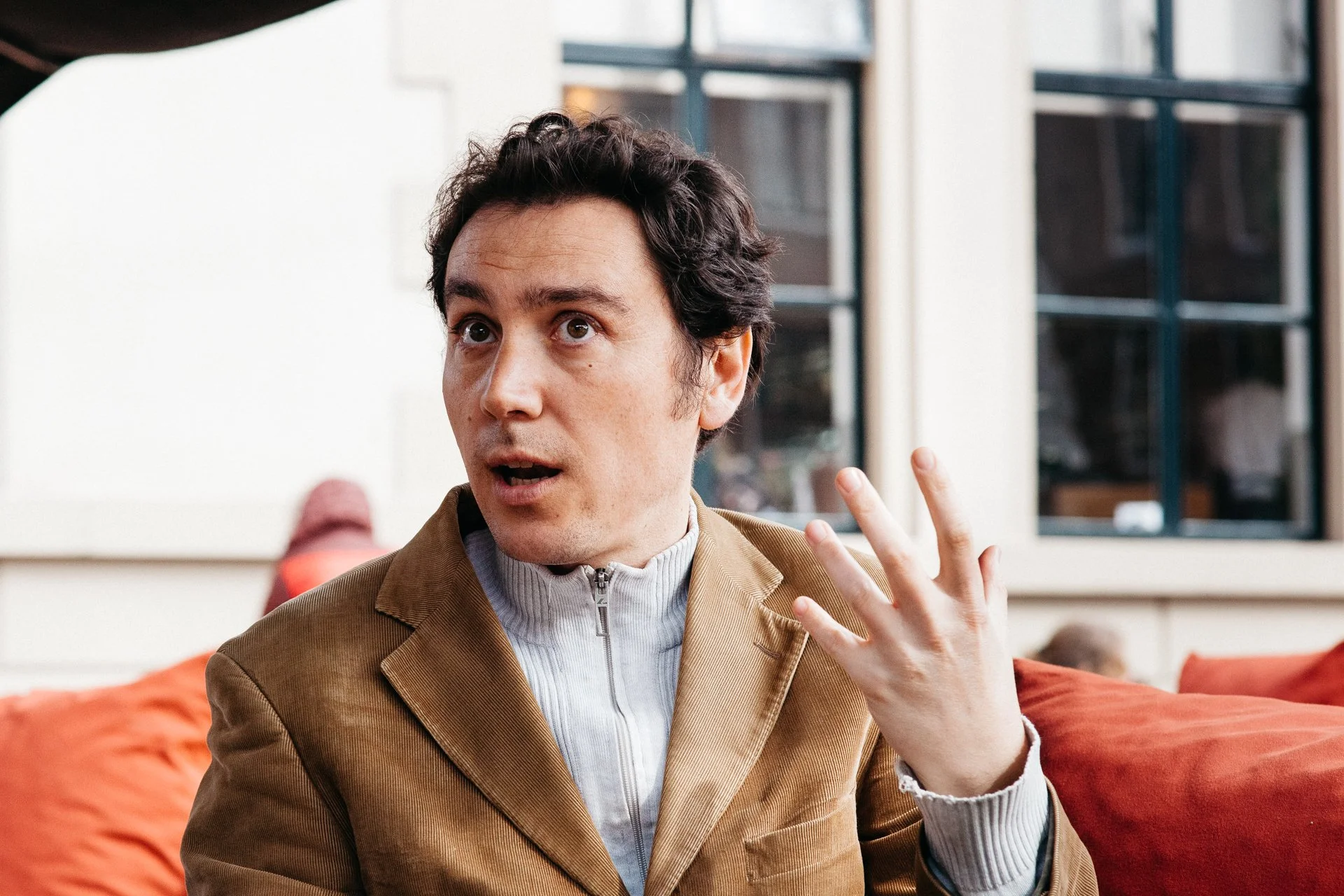 A man with dark curly hair and fair skin gestures with his right hand while seated on an orange cushion in a cafe or restaurant, with large windows in the background.