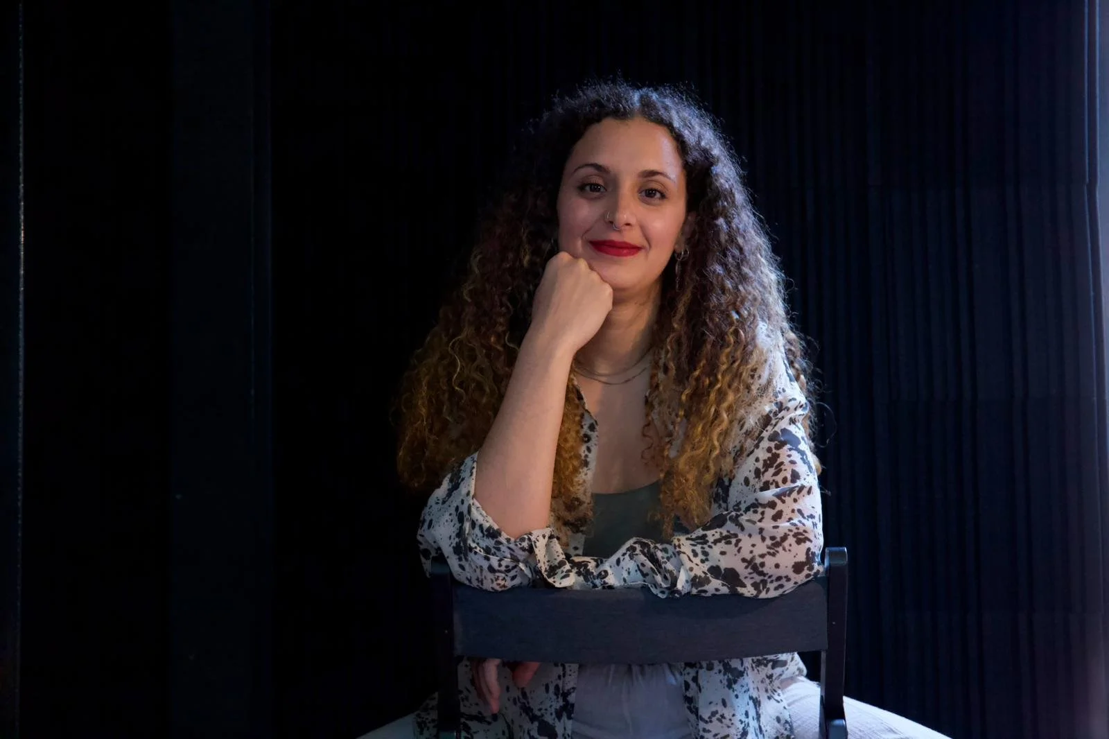 A woman with curly hair sitting on a chair, resting her chin on her fist, smiling, with a dark curtain background and light coming from the right side.