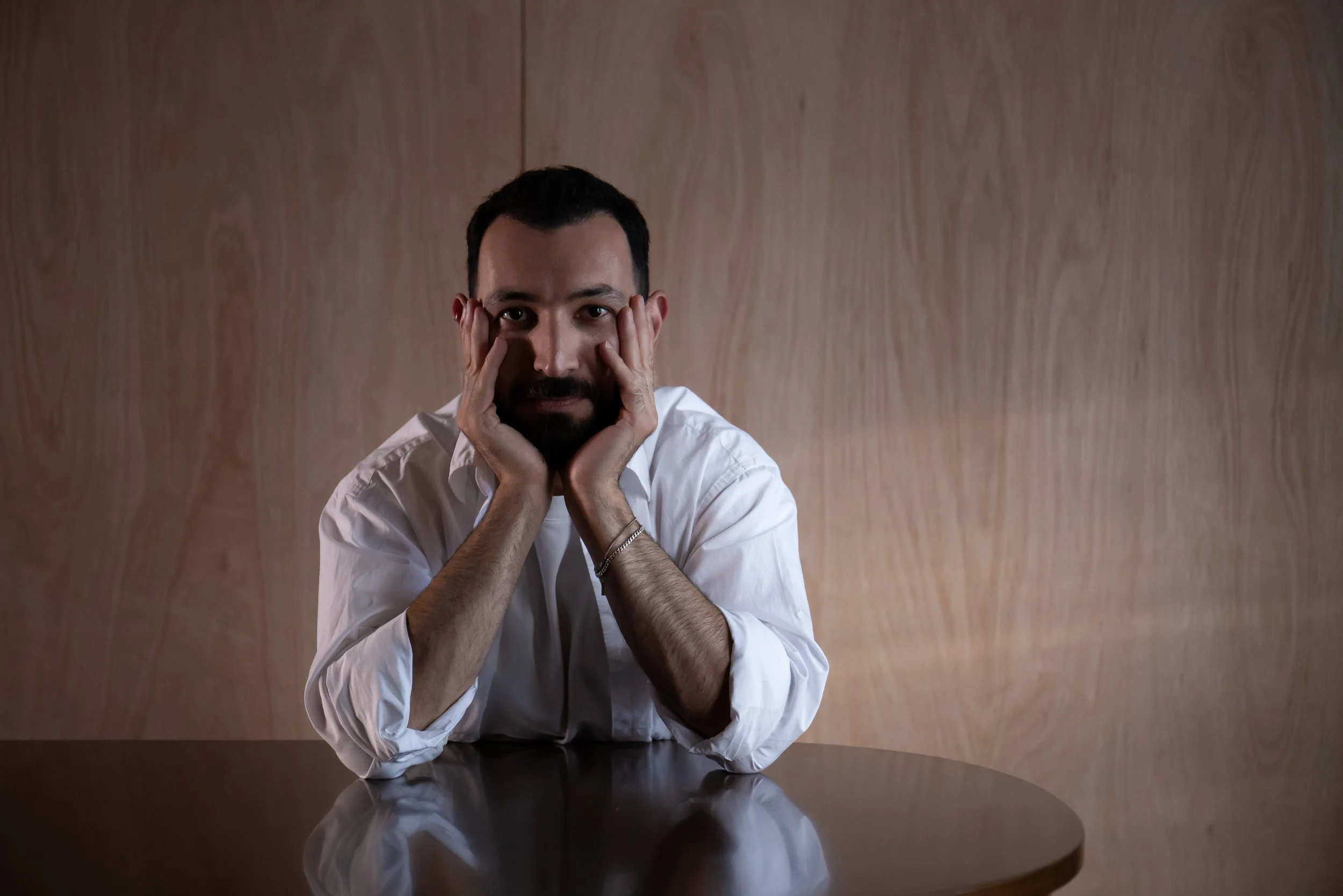 A man with dark hair, beard, and light skin is sitting at a round table with a dark surface, resting his head on his hands, with elbows on the table. He is wearing a white shirt and looking directly at the camera with a neutral expression. The background is a plain, wooden wall.