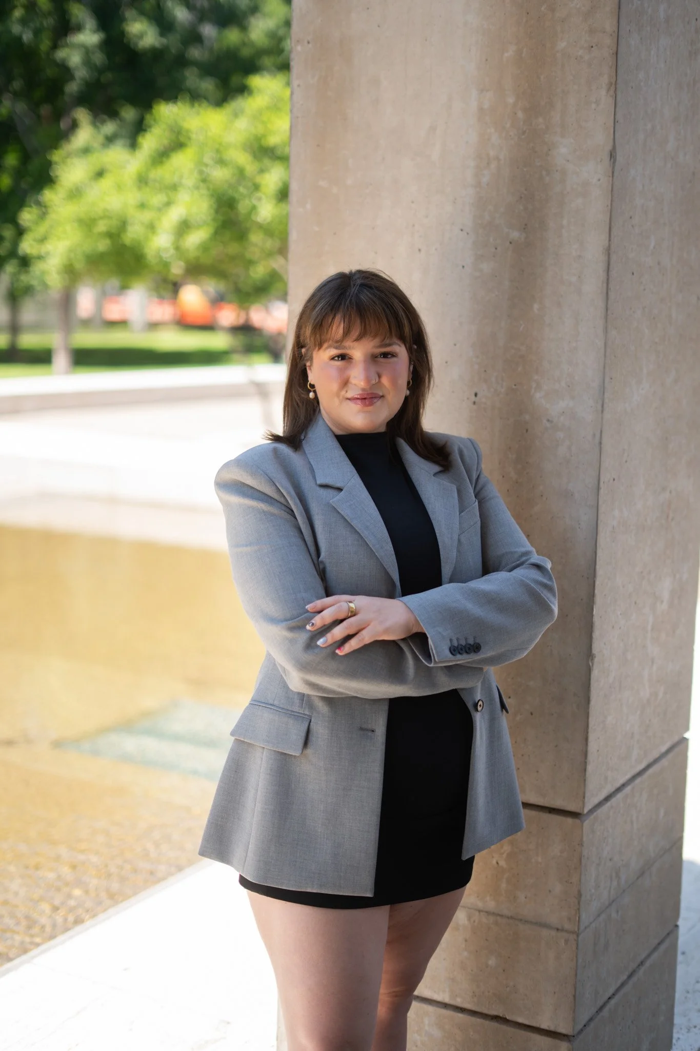 Ashley Goodwin wearing a grey blazer and black dress standing outdoors next to a concrete pillar with arms crossed, with green trees and a water feature in the background.