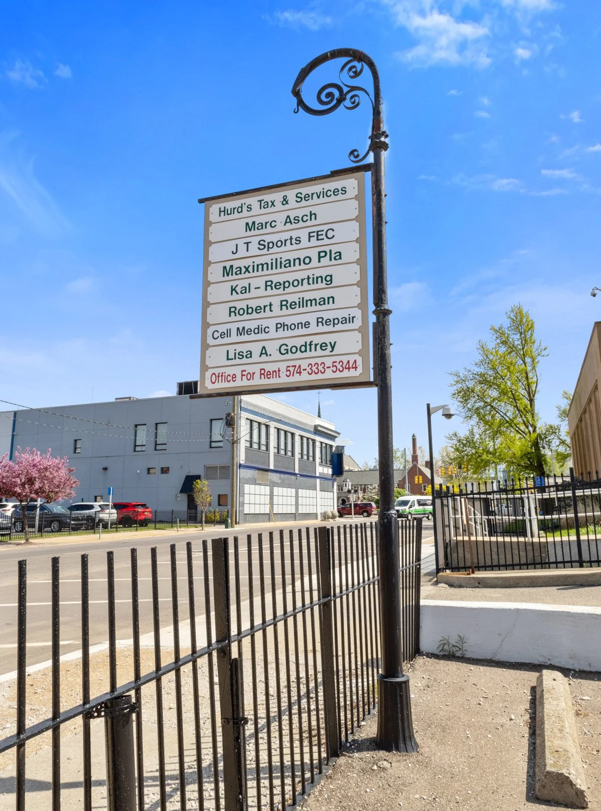 A street view with a tall decorative signpost listing various local businesses and an office space for rent, set against a clear blue sky and some trees.