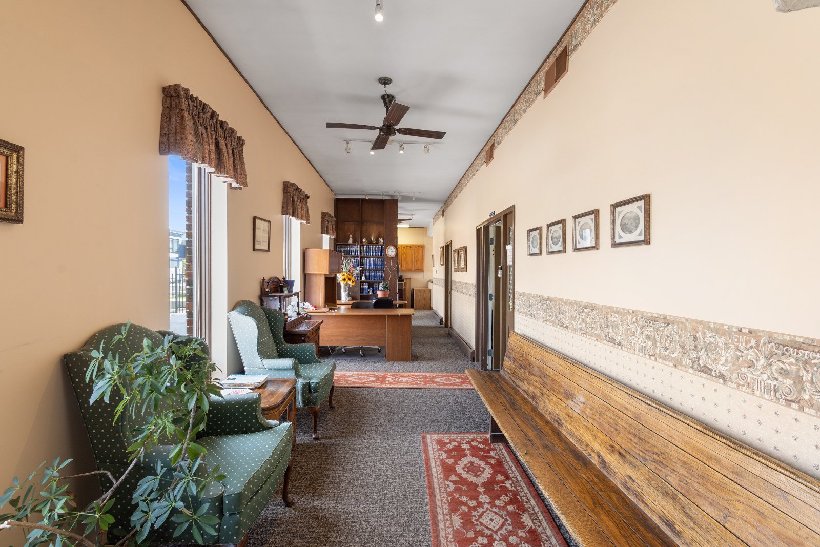 Interior of a waiting area or reception in a building, with upholstered armchairs, a wooden bench, a small table, and a desk at the far end. There are framed pictures on the wall, a ceiling fan, and large windows with curtains.