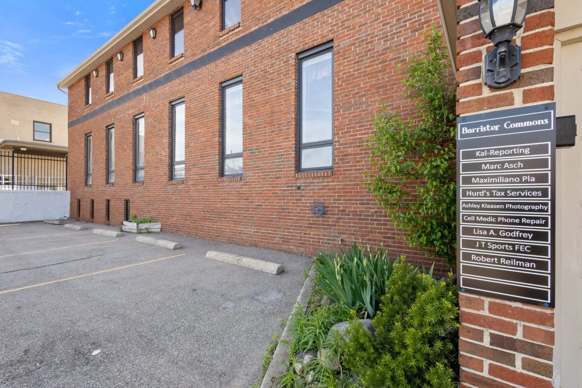 Exterior view of a brick building with a parking lot in front. There is a sign listing business names next to the building, including Barrister Commons, Kal-Reporting, Marc Asch, Maximiliano Pla, Hurd's Tax Services, Ashley Klaasen Photography, Cell Medic Phone Repair, Lisa A. Godfrey, J T Sports FEC, and Robert Reilman.