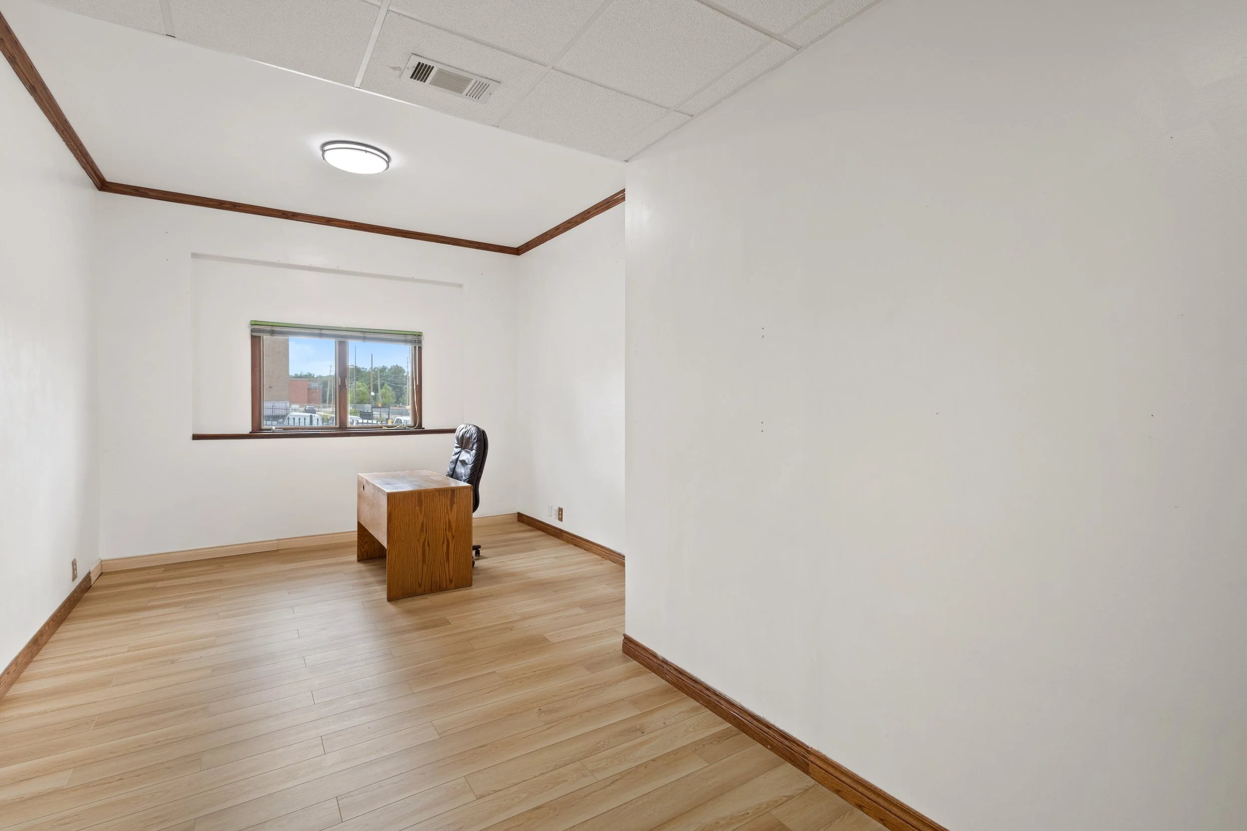 An empty office room with white walls, a window, a wooden desk, and a black office chair.