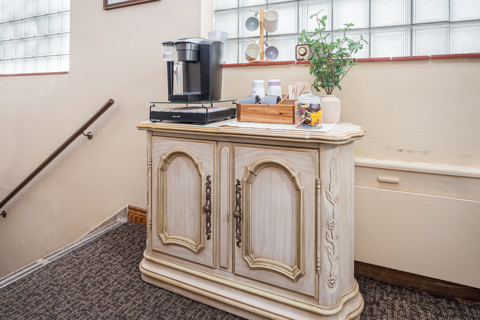 A vintage style wooden cabinet with ornate doors holds a coffee station with a coffee machine, cups, and snacks, next to a plant and glass block wall.