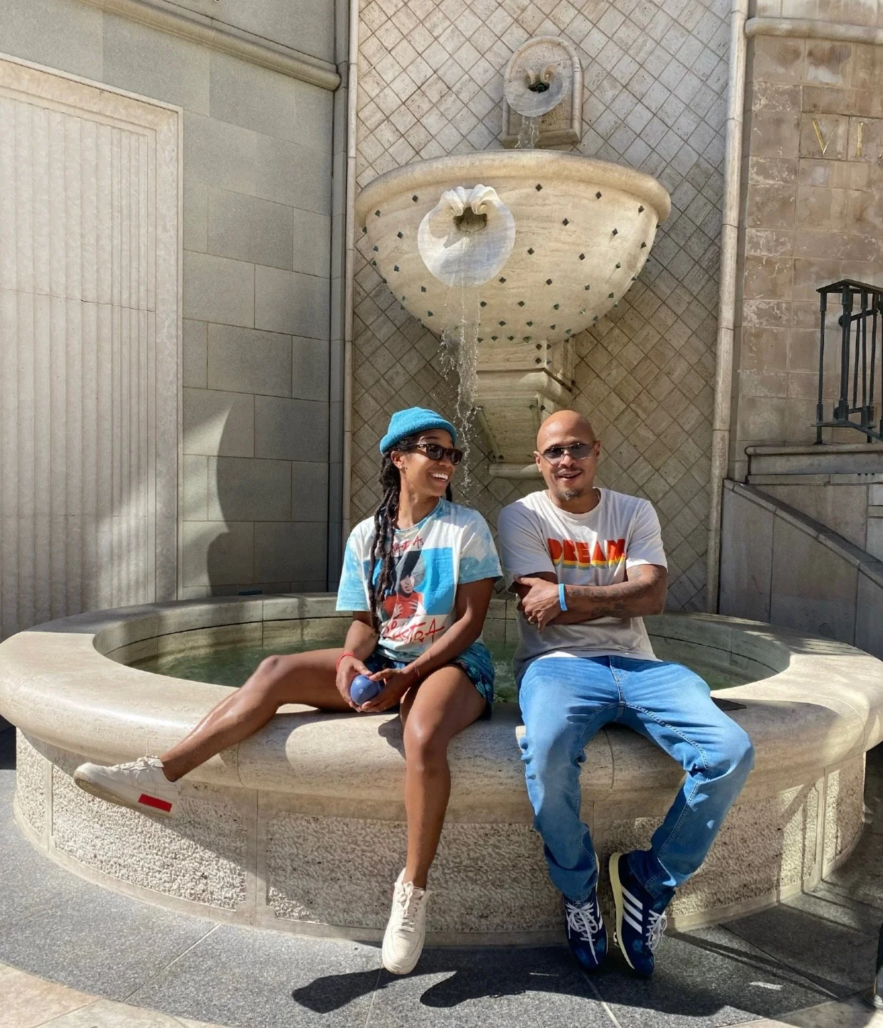 Two people sitting on the edge of a fountain with water flowing from a decorative wall-mounted spout. The woman is wearing a blue beanie, sunglasses, a graphic t-shirt, shorts, and white sneakers. The man is wearing sunglasses, a white t-shirt with colorful text, jeans, and Adidas sneakers. They are smiling and enjoying a sunny day.