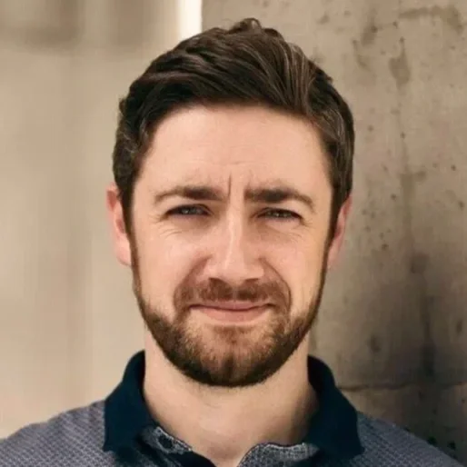 A man with short brown hair and a beard, standing outdoors near a concrete wall, looking at the camera with a slight smile.