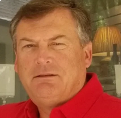 A middle-aged man with light brown hair and tan skin wearing a red collared shirt, winking with a serious expression.