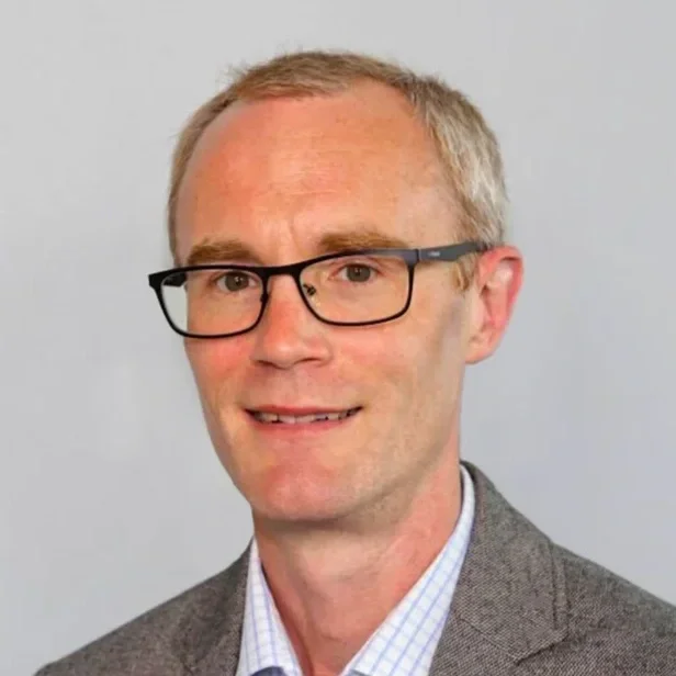 A man with short blond hair, glasses, wearing a gray blazer and collared shirt, smiling at the camera.