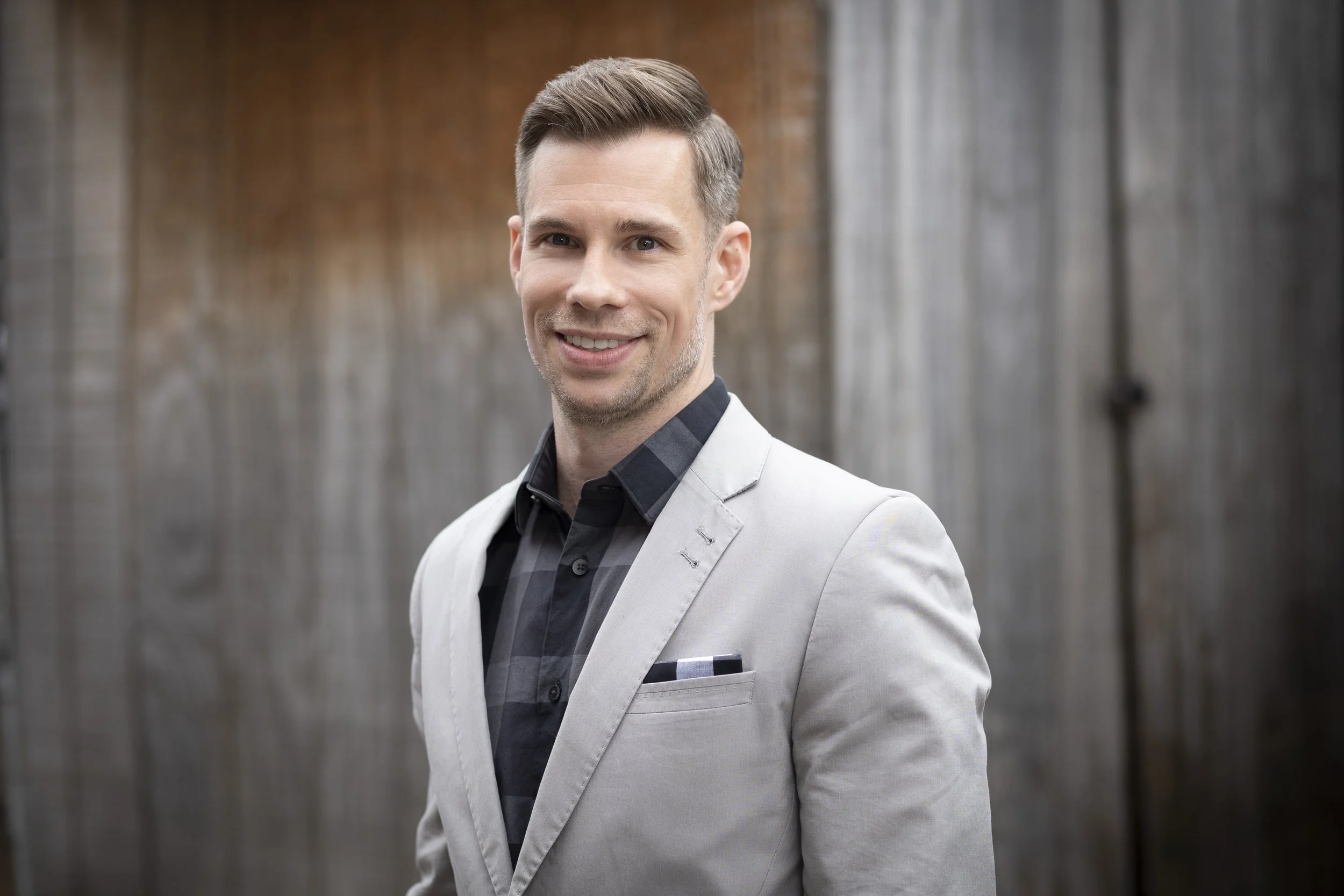 A smiling man wearing a light gray blazer over a dark plaid shirt, standing outdoors in front of a wooden panel background.