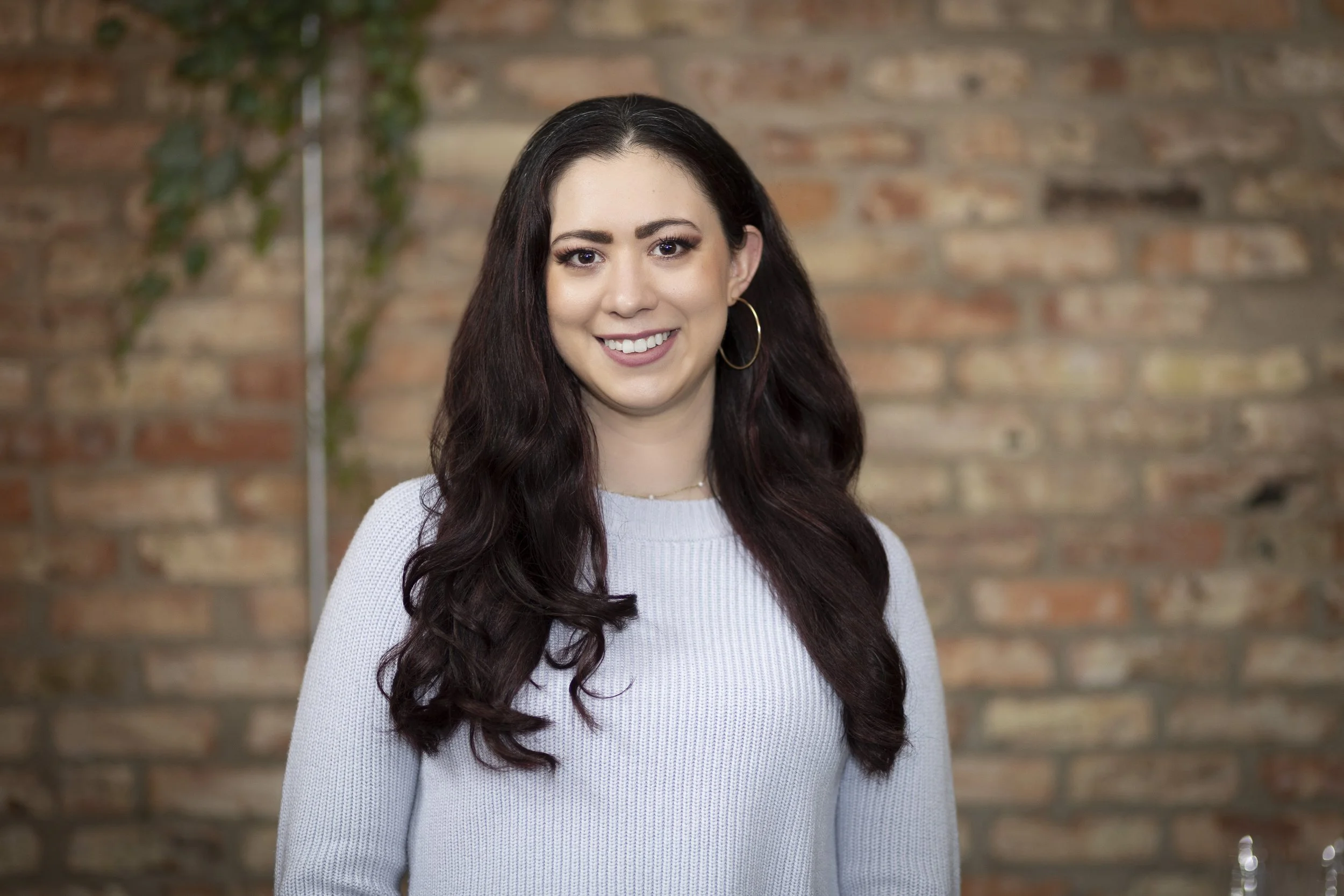 Portrait of a young woman with long dark hair, smiling, wearing a light gray sweater, gold hoop earrings, standing in front of a brick wall with a plant to her left.