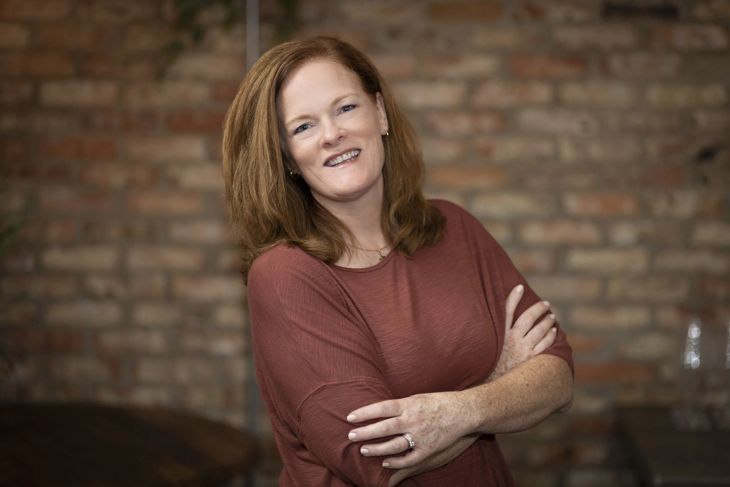 A woman with red hair, wearing a rust-colored top, standing with her arms crossed in front of a brick wall, smiling.