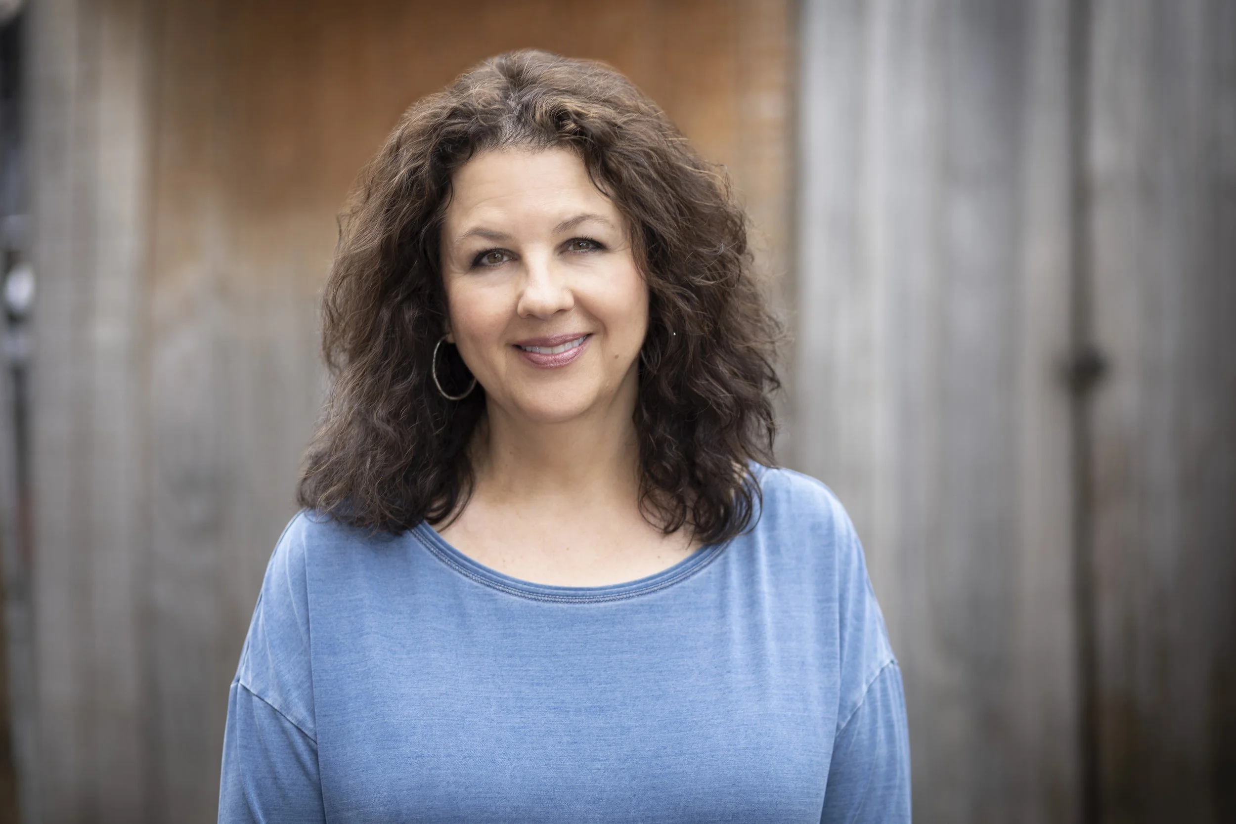 A woman with curly brown hair wearing a blue top, standing outdoors with a blurred wooden fence in the background.