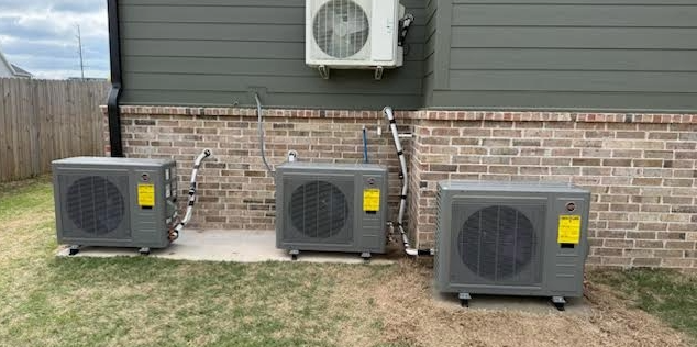 Three air conditioning outdoor units installed on the ground outside a house with brick and siding exterior