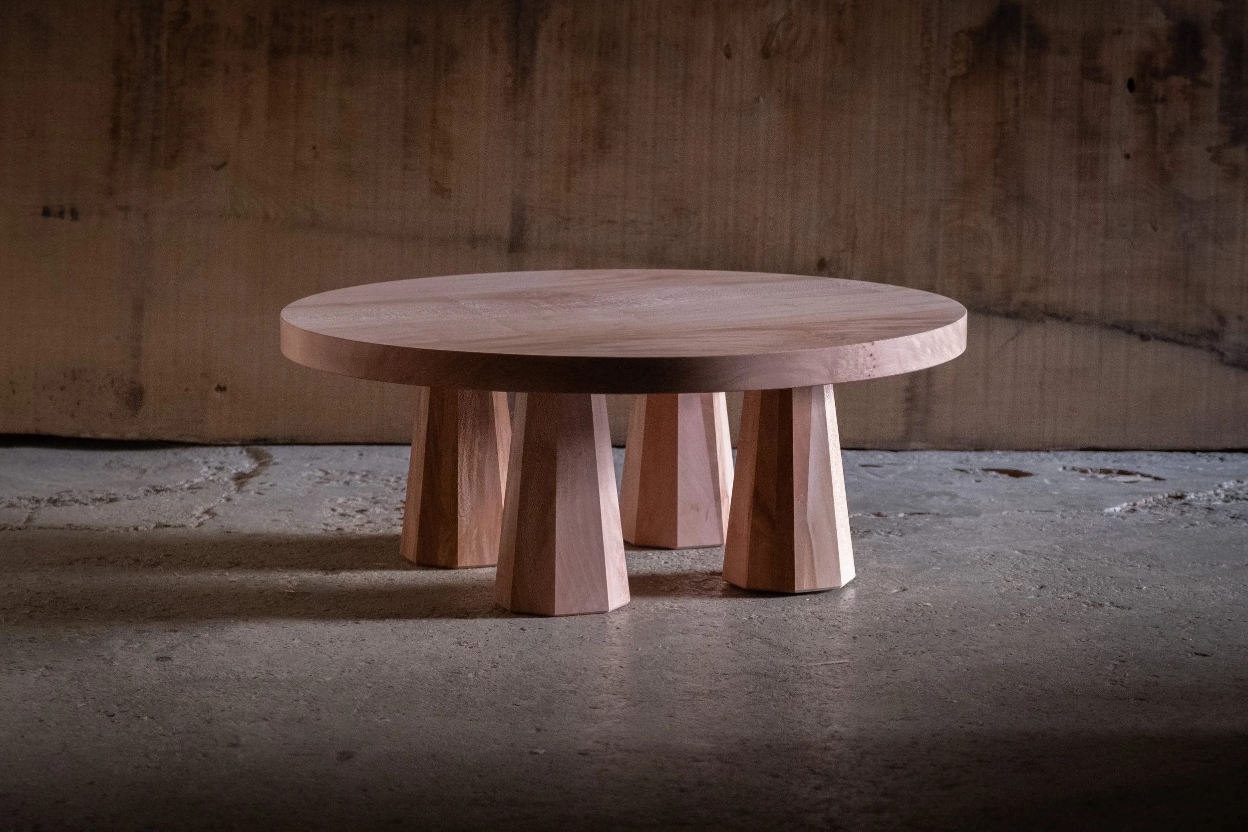 A round wooden table with short, thick legs on a concrete floor against a wooden background.