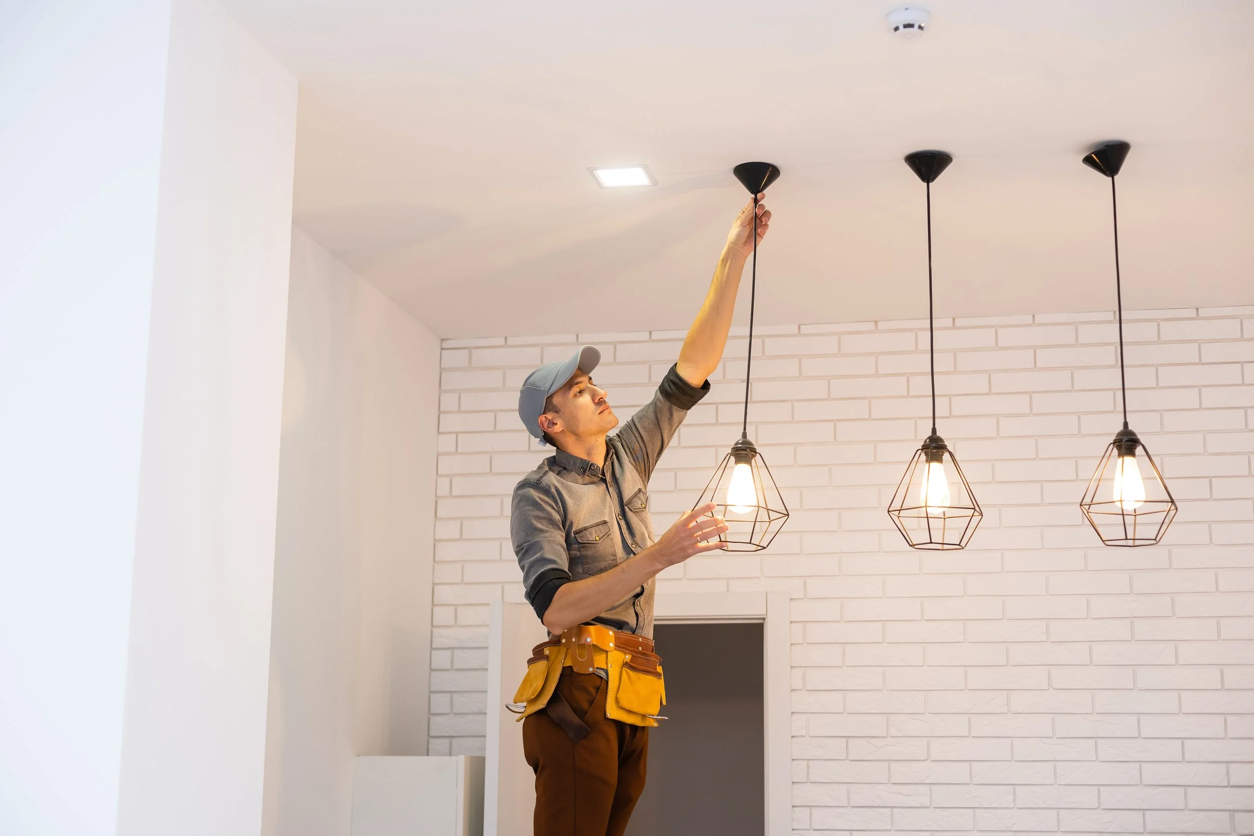 Electrician adjusting pendant light on ceiling in modern room with brick wall.