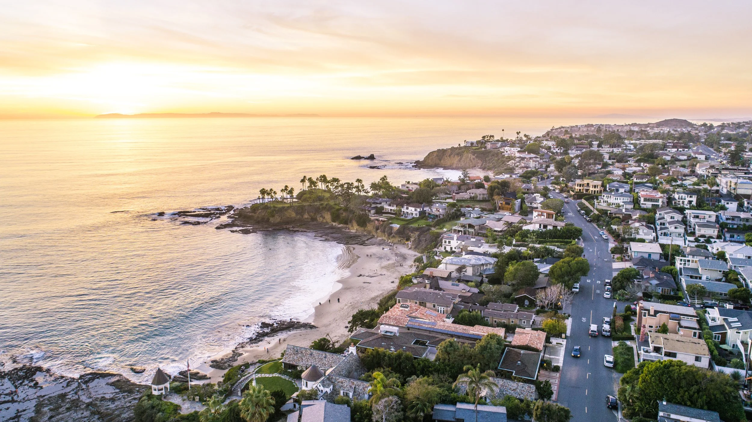 Aerial view of a coastal town at sunset with houses, a beach, and palm trees.