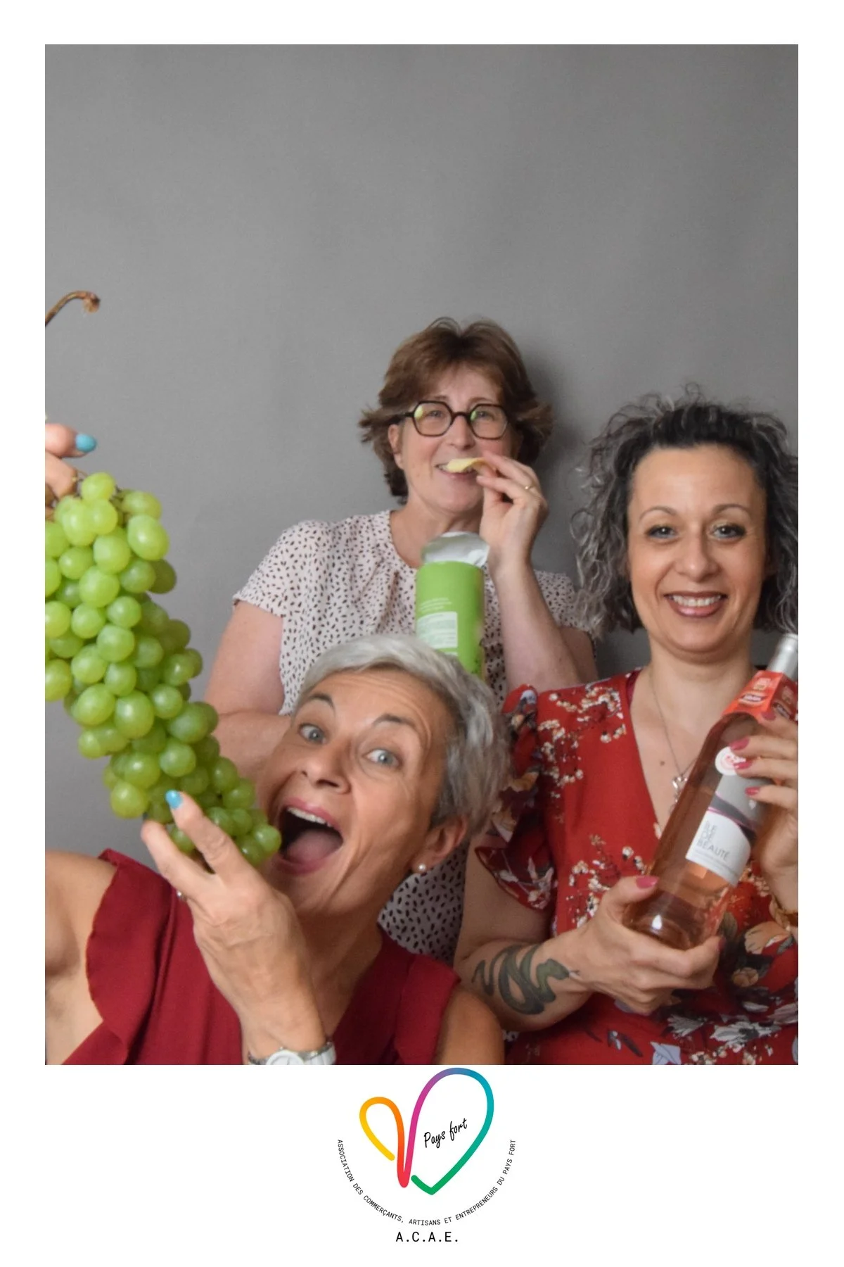 Groupe de trois femmes souriantes posant avec du vin, des raisins verts et des snacks, devant un fond gris, semble lors d'une fête ou d'un rassemblement.