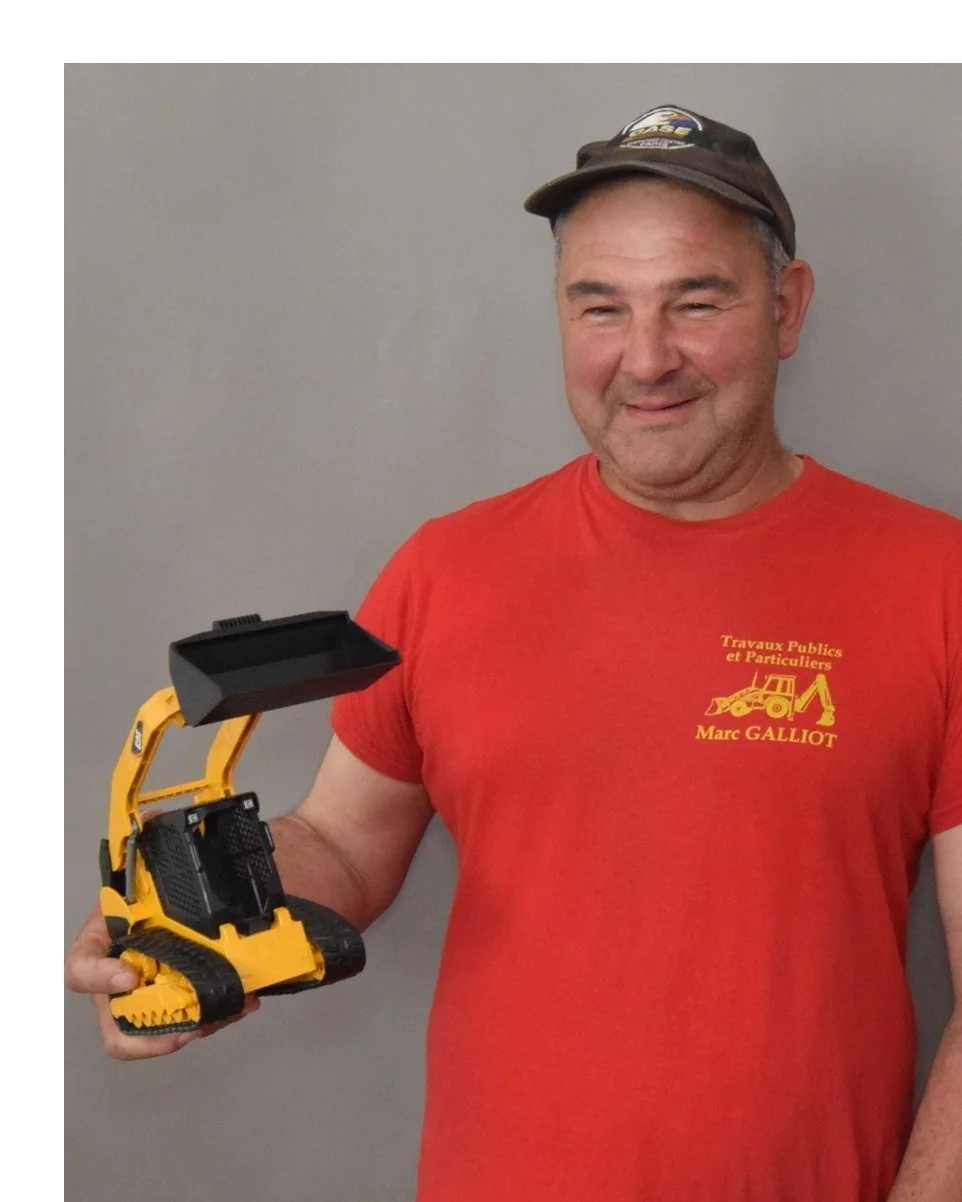 Homme souriant portant un t-shirt rouge avec l'inscription en français, un logo de pelle, et le nom « Marc Galliot » ; il tient une petite pelleteuse télécommandée jaune et noire contre un mur gris.