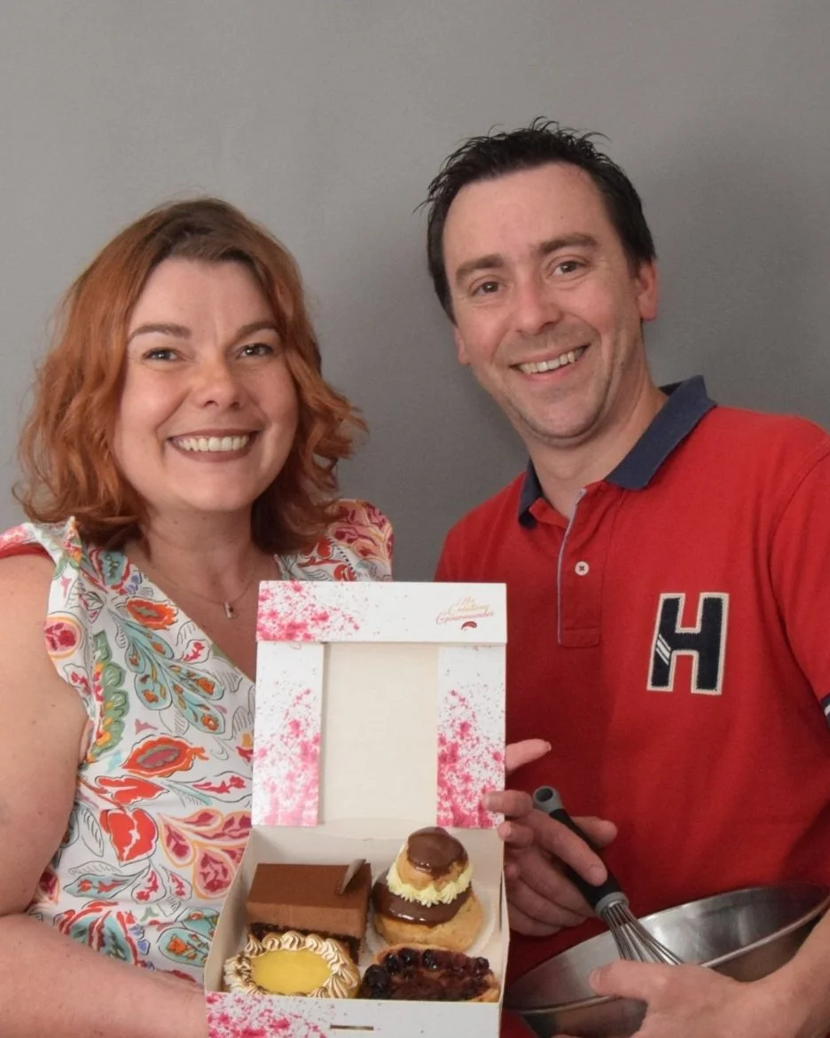 Un homme et une femme souriants posent avec une boîte de pâtisseries. La femme tient la boîte ouverte, affichant plusieurs desserts, et l'homme tient un fouet.