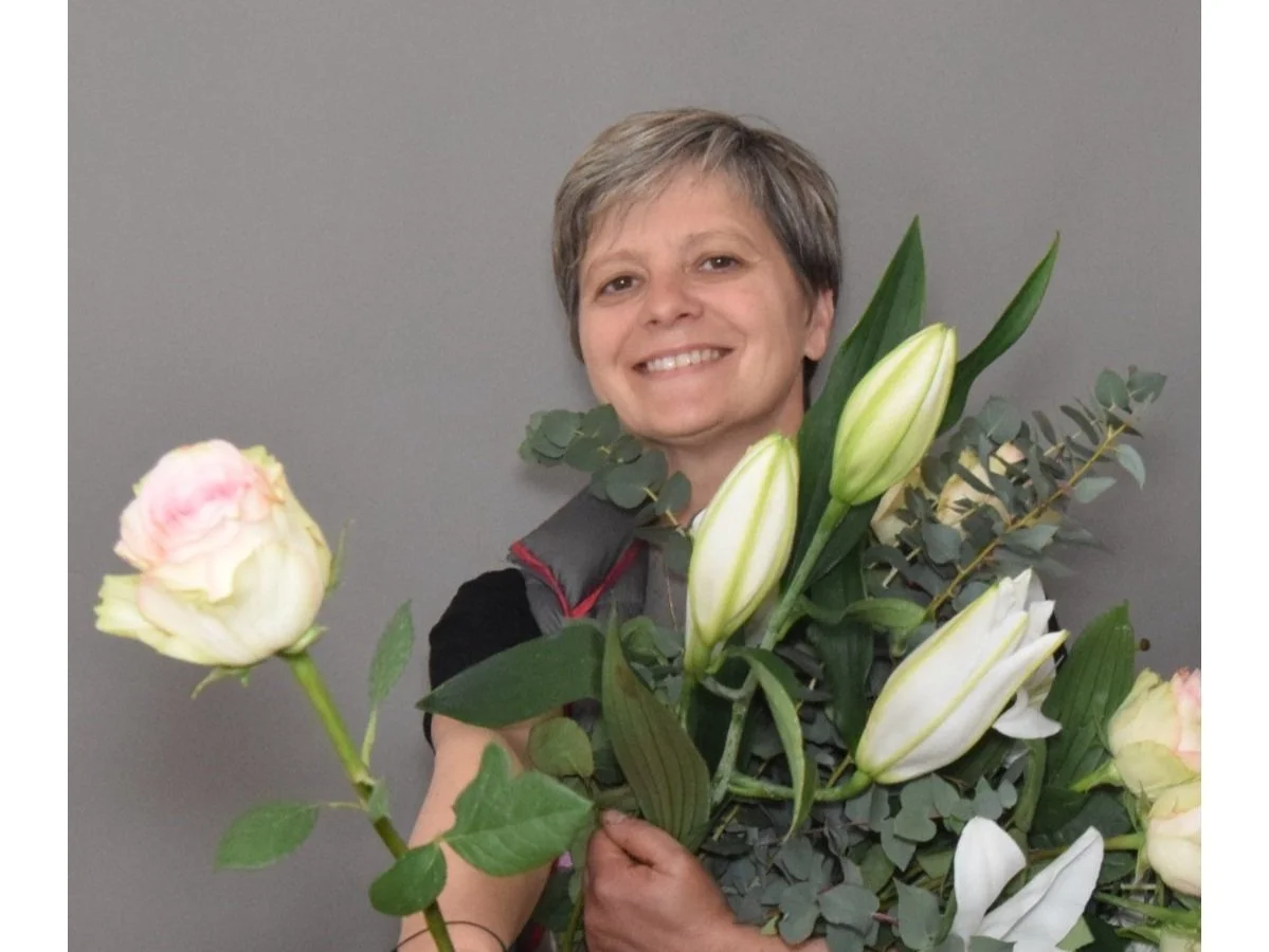 Une femme souriante tient un grand bouquet de fleurs, comprenant principalement des lys blancs et quelques pivoines roses pâles, contre un fond gris.