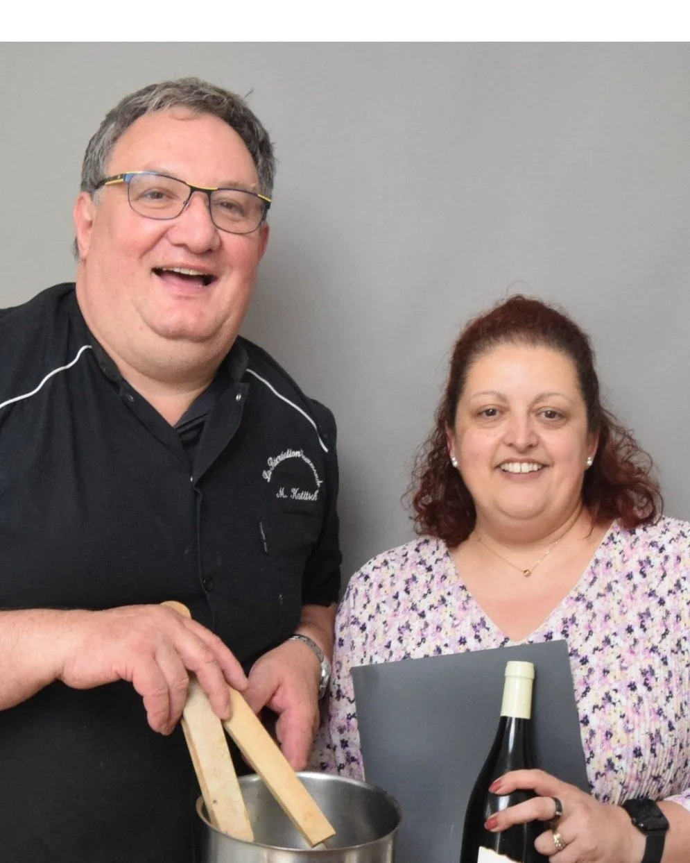 Un homme et une femme souriants posant ensemble, l'homme tenant une cuillère en bois dans un pot et la femme tenant une bouteille de vin, contre un mur gris.
