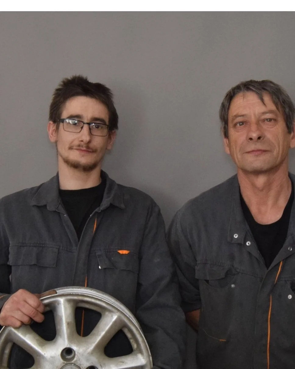 Deux hommes posent devant un mur gris, l'homme de gauche tient une pièce de machine en métal.