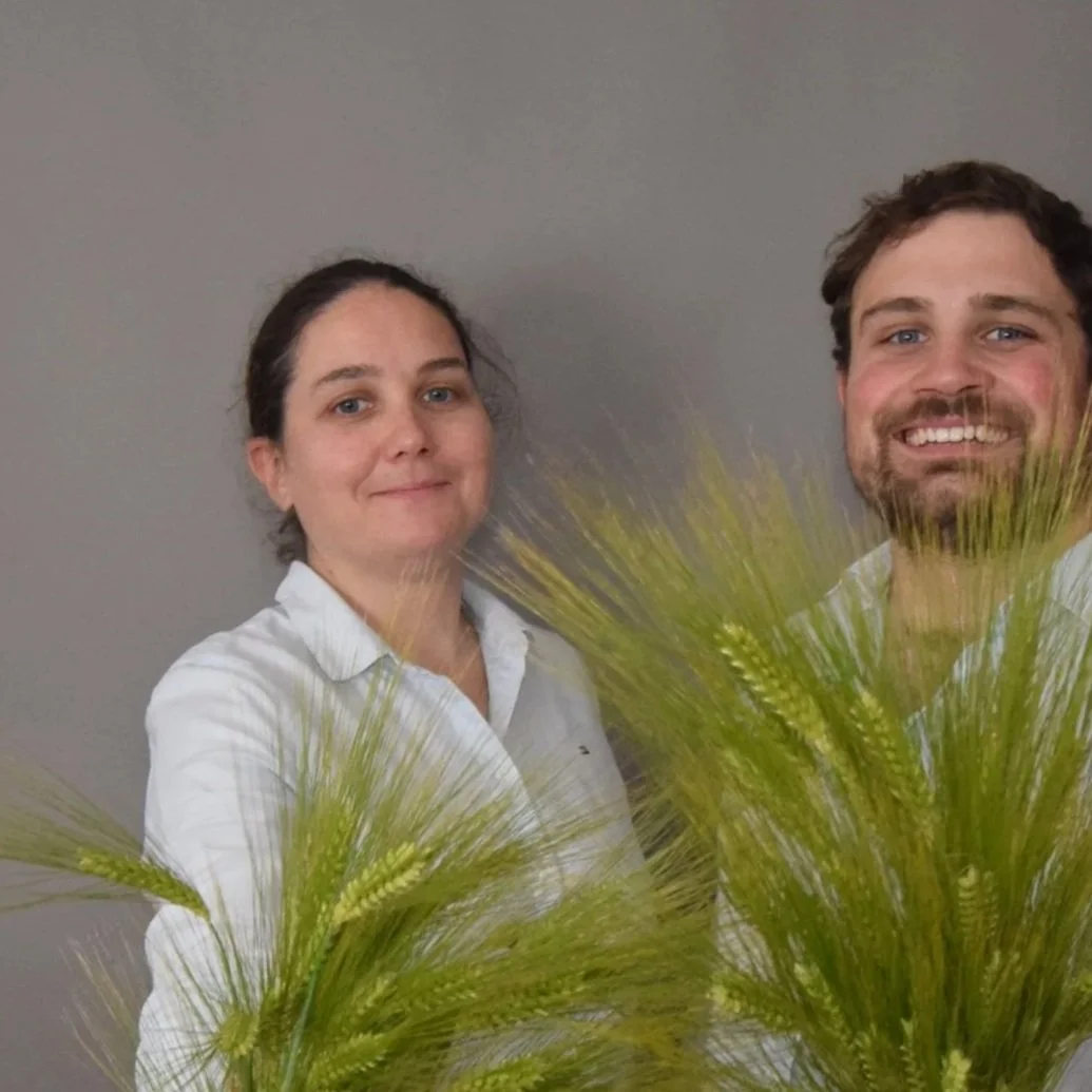 Deux personnes posent avec des plantes de céréales devant un mur gris.