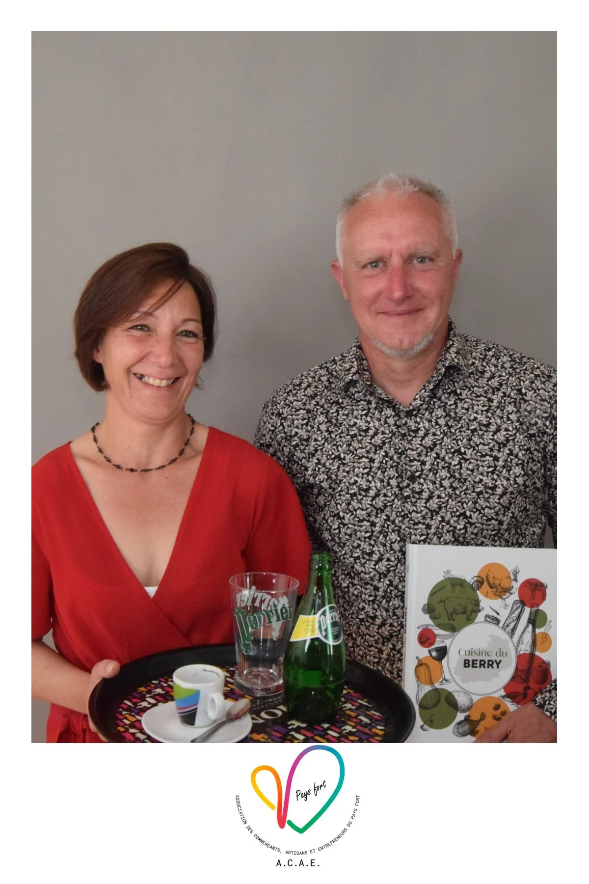 Deux personnes, un homme et une femme, posent ensemble, souriants devant un mur gris. La femme porte une robe rouge et une chaîne noire, l'homme porte une chemise à motifs floraux. They tiennent un plateau avec une tasse, une bouteille de Perrier, un