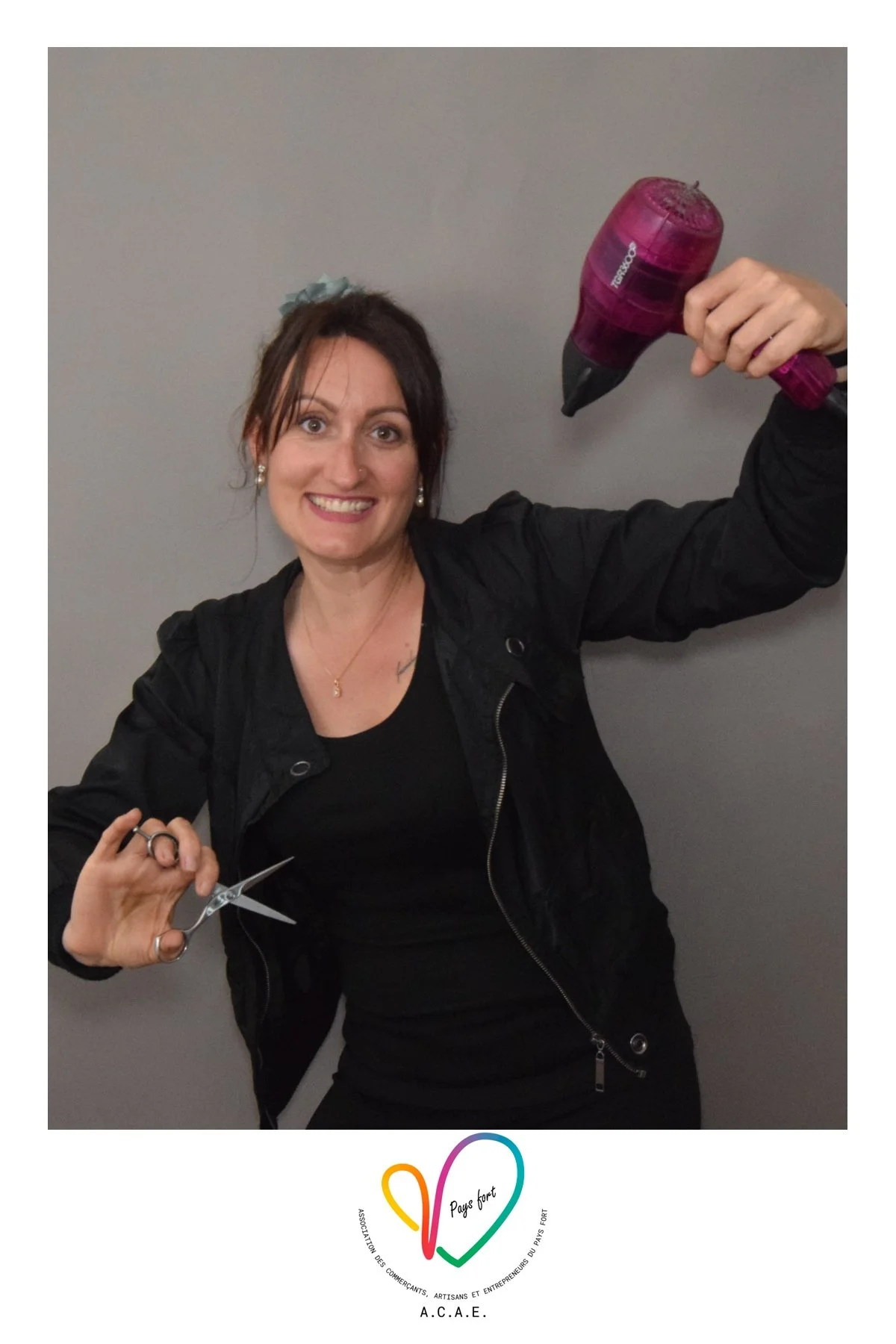 Une femme souriante tient une paire de ciseaux dans une main et un sèche-cheveux dans l'autre. Elle porte un blazer noir et une chemise noire, avec un fond gris. Il y a un logo en bas de l'image avec le texte "Pays Fort" et d'autres inscriptions.