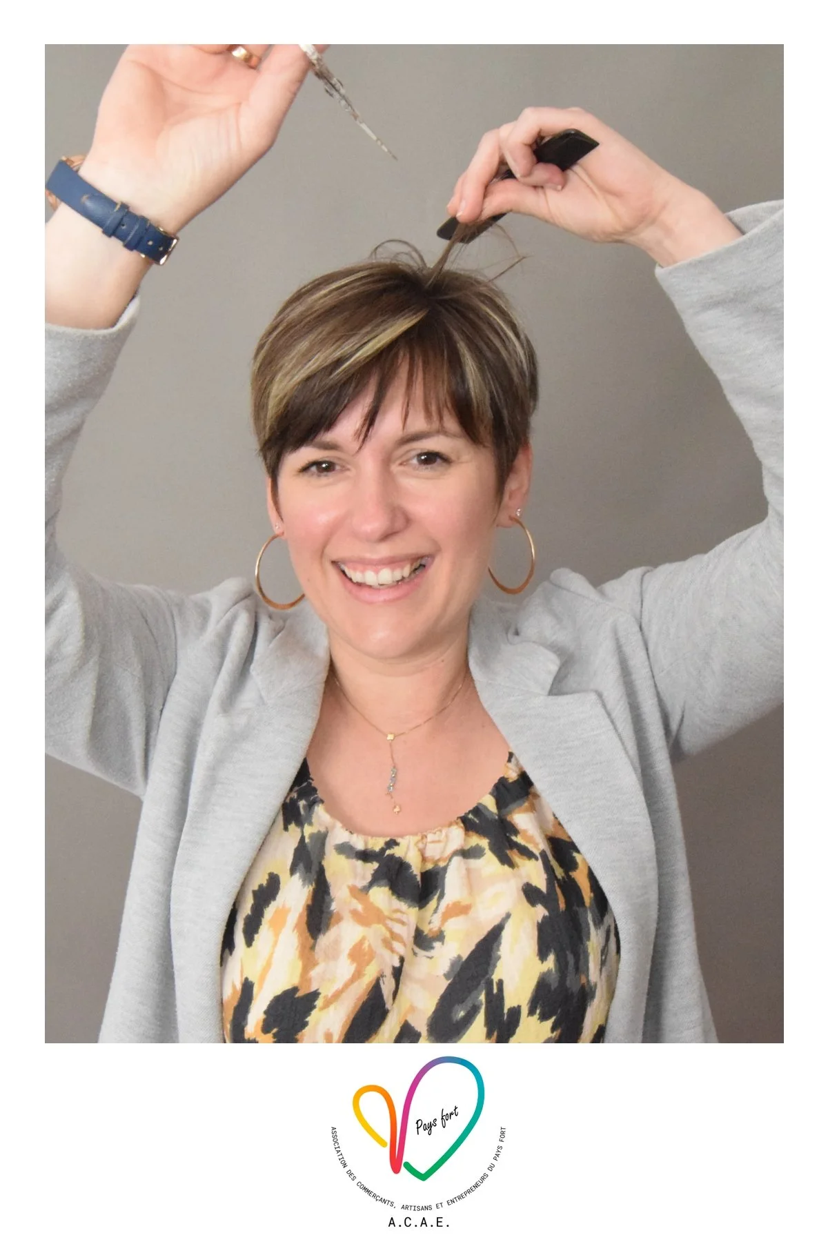 Une femme souriante avec une coupe de cheveux courte et brune, portant des boucles d'oreilles en hoop, un collier et un blazer gris, tient une aiguille et une paire de ciseaux, posant devant un fond gris, avec un logo coloré en forme de cœur en bas d