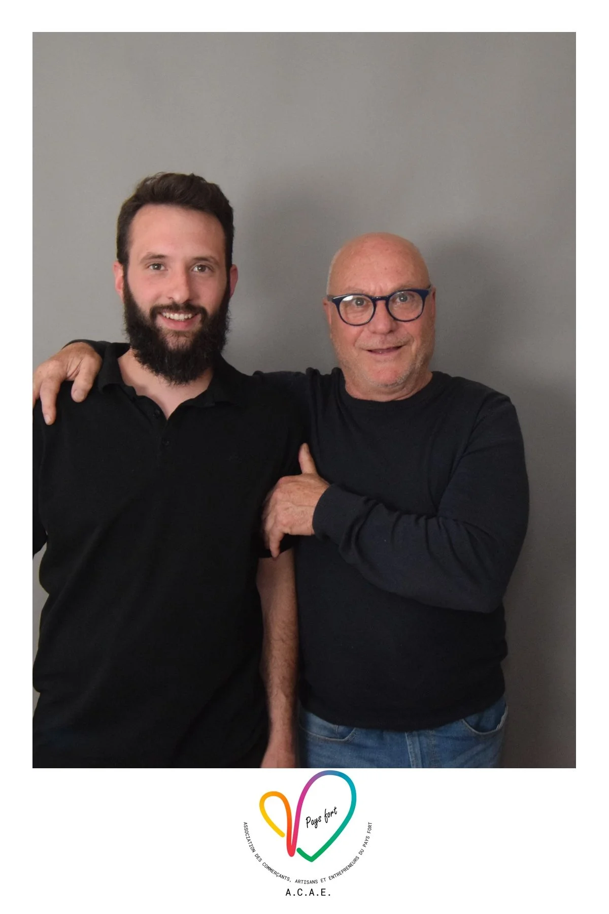 Deux hommes souriants, l'un avec une barbe et l'autre avec des lunettes, posent ensemble devant un mur gris. Ils portent tous deux des vêtements noirs. Il y a un logo en bas avec un cœur coloré et le texte « Pays Fort » et « A.C.A.E. ».