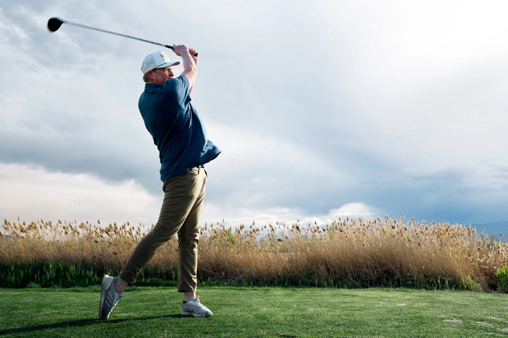 Man in a blue shirt and khaki pants swinging a golf club on a golf course with cloudy sky and tall grasses in the background.