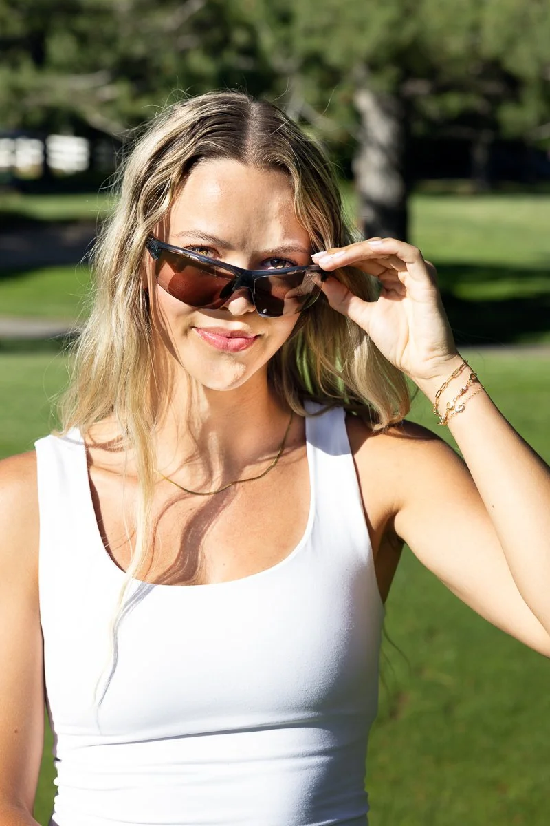 Young woman with long blonde hair wearing sunglasses and a white tank top outdoors on a sunny day.