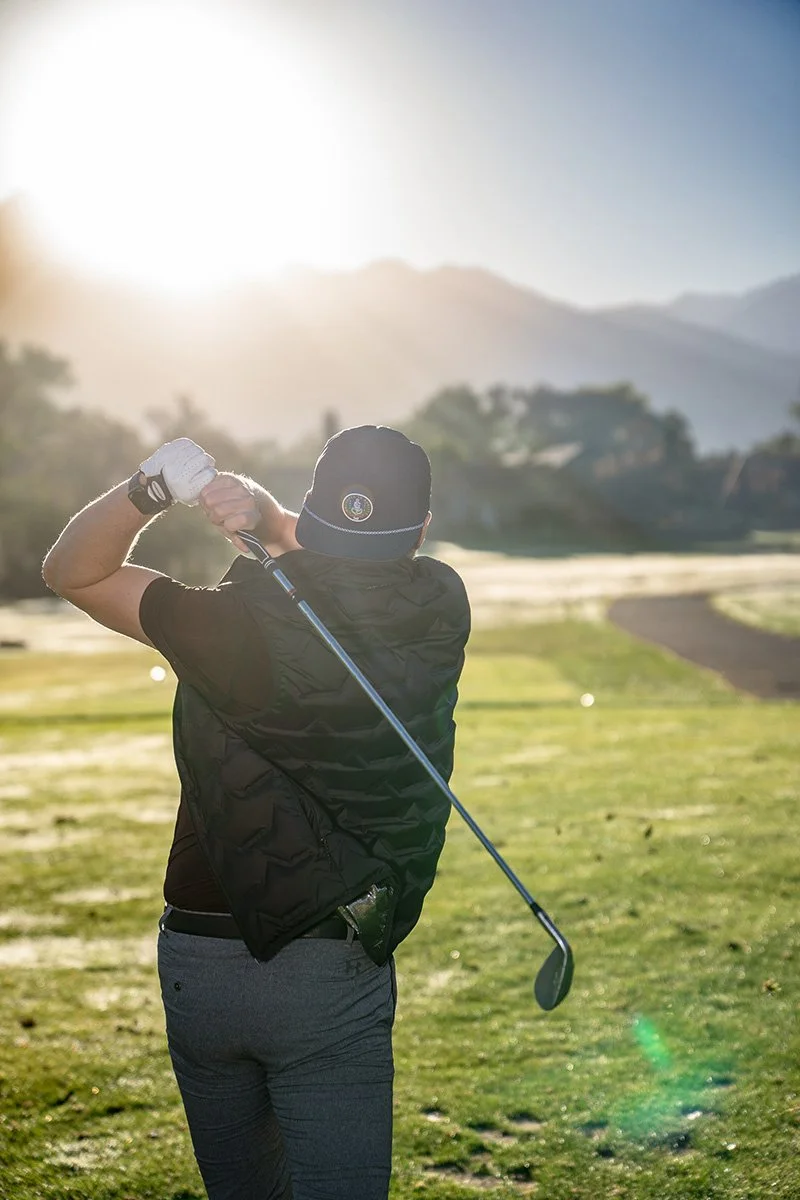 A person playing golf on a course during sunrise or sunset, swinging a golf club with mountains in the background.