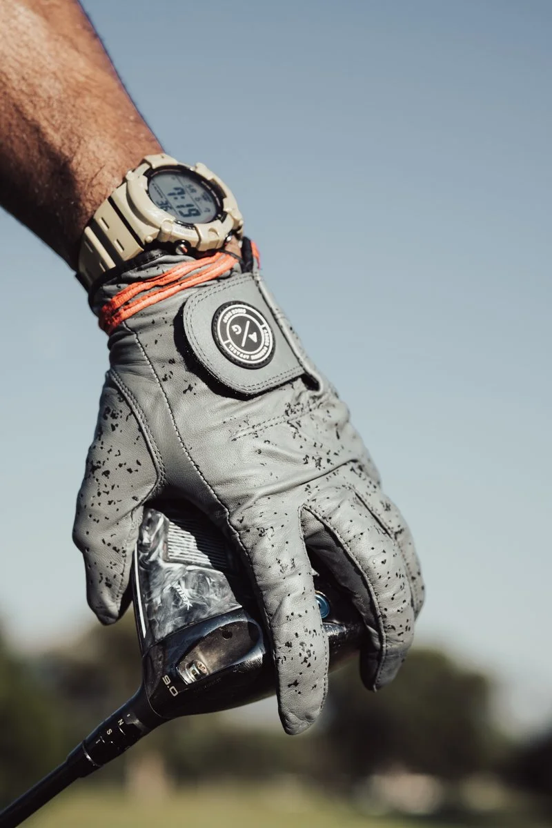 A close-up of a person's hand wearing a grey golf glove holding a golf club, with a beige sports watch on their wrist and an orange bracelet, against a clear blue sky.