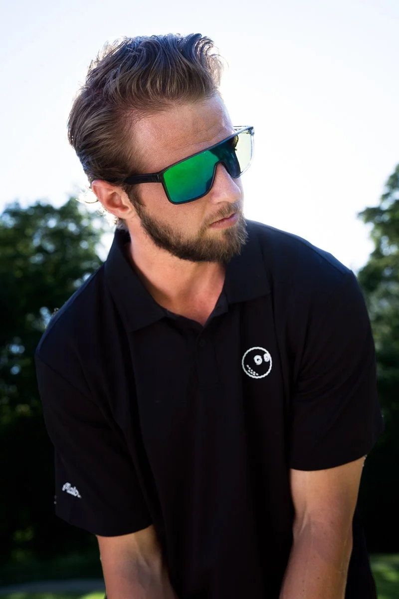 A man with styled hair and a beard wearing large reflective sunglasses and a black polo shirt with a small white smiling face logo, outdoors with trees and a bright sky in the background.
