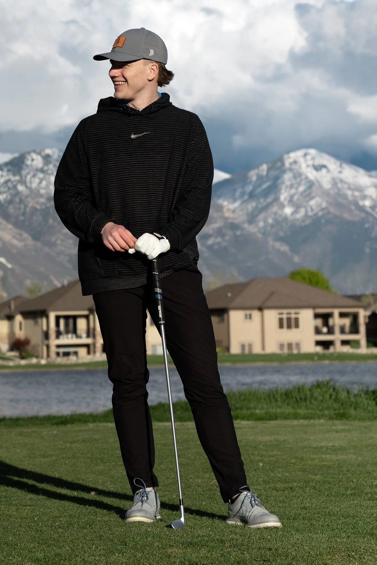 A young man smiling while standing on a golf course with mountains in the background, holding a golf club.