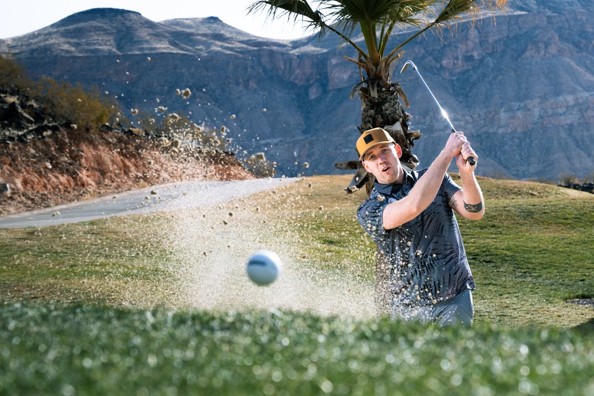 A young man hitting a golf ball out of a sand trap on a golf course, with mountains and a palm tree in the background.
