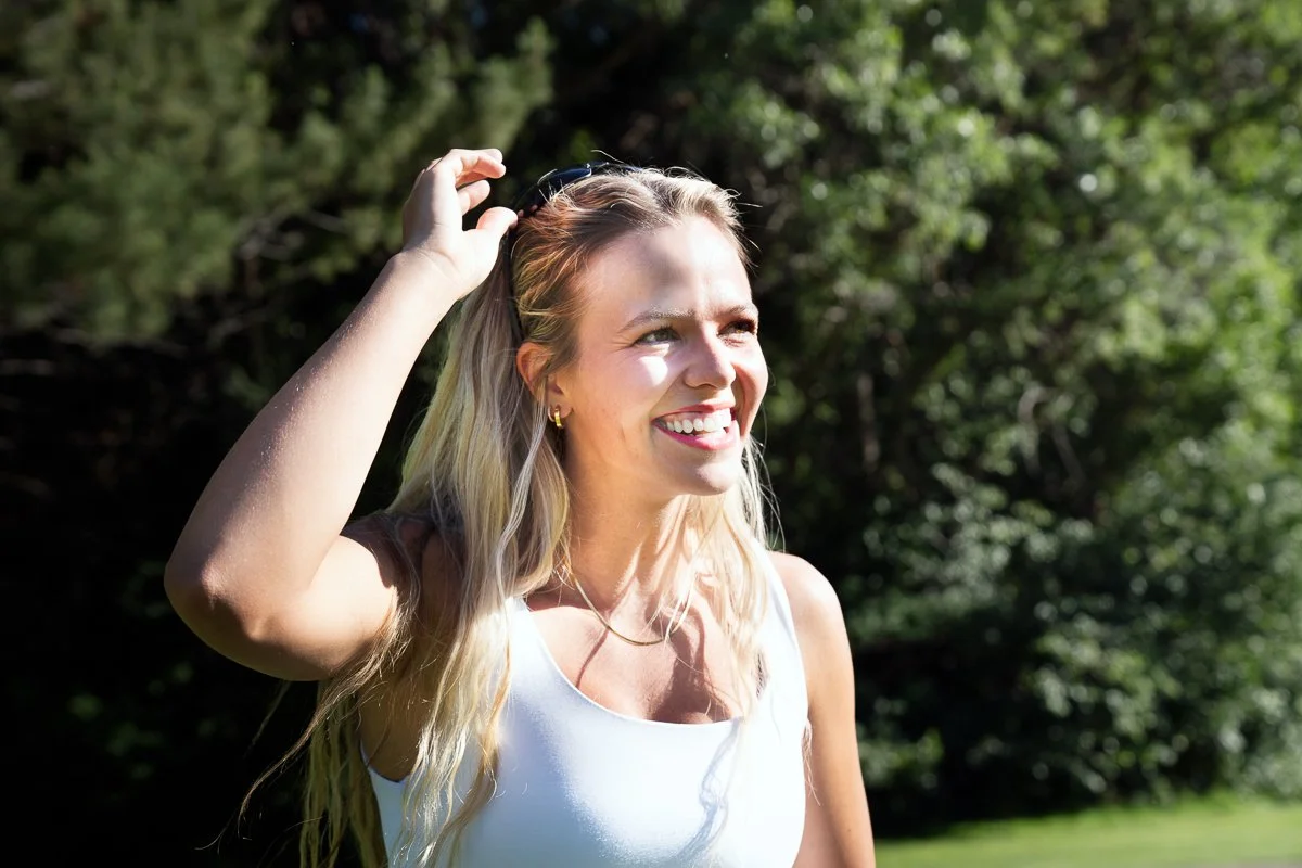 A young woman with blonde hair smiling outdoors on a sunny day, holding sunglasses on her head with trees in the background.