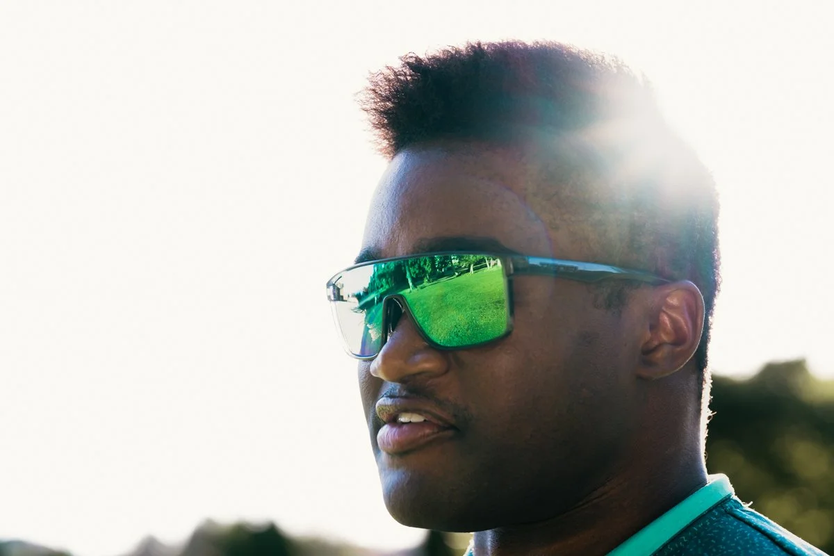 Close-up of a young man wearing reflective sunglasses outdoors with sunlight shining from behind, and trees in the background.